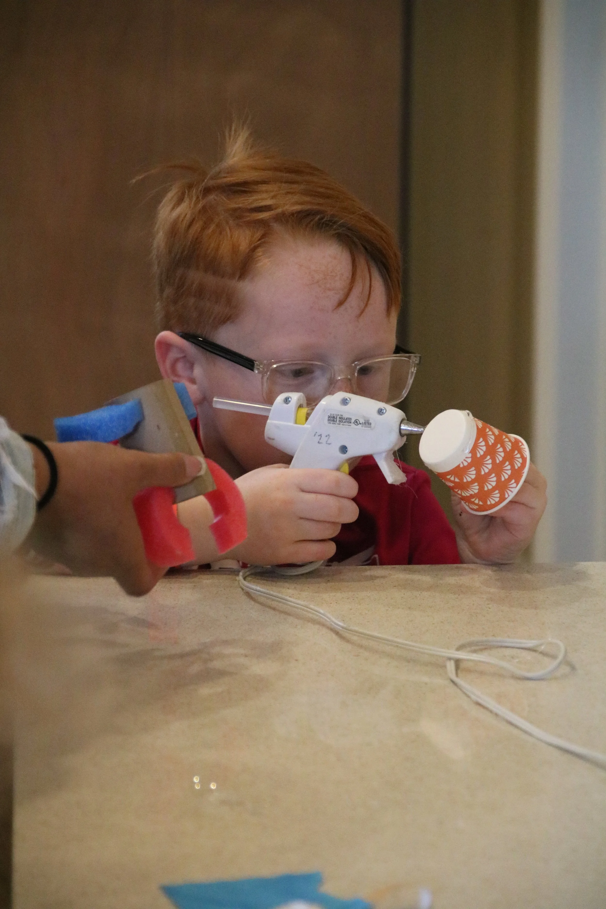 A young boy with red hair and glasses focusing on a small, orange paper cup using a glue gun, with an adult's hand guiding him, on a table.