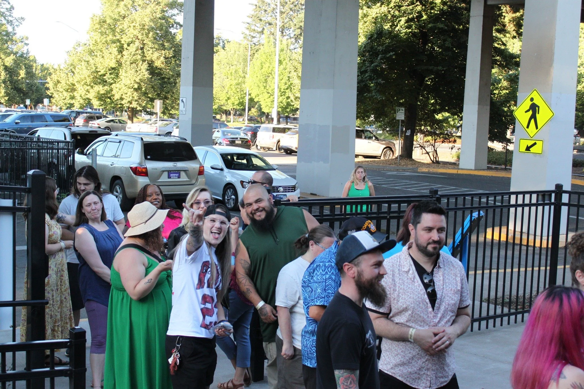 Group of people waiting outdoors near a parking lot, some smiling and talking, under a bridge with trees in the background.