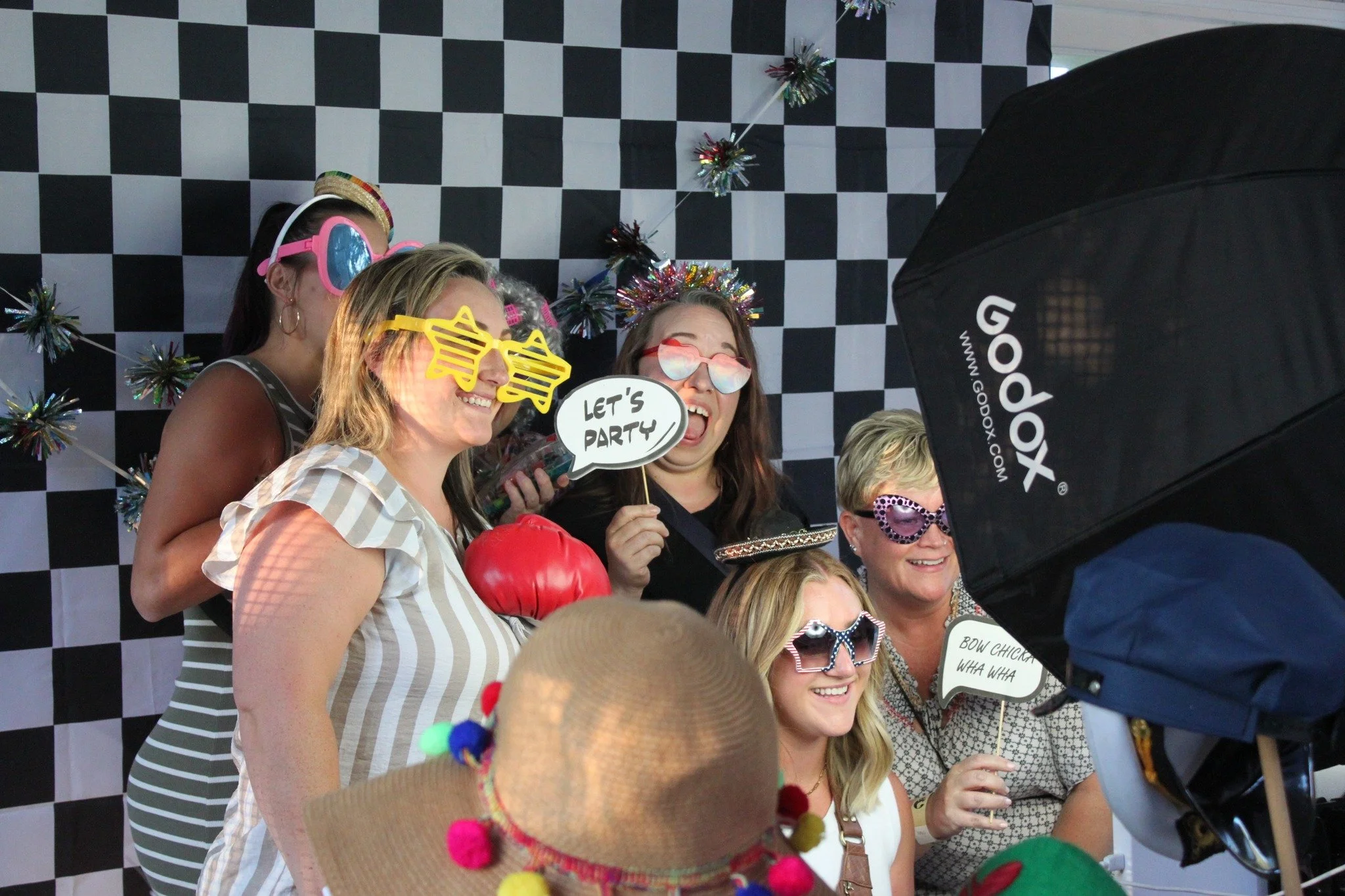 Group of women at a party wearing colorful costumes, novelty glasses, and holding playful signs, with a checkered backdrop and party decorations.