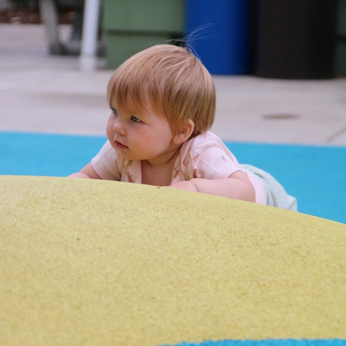 A young child with red hair lying on their stomach on colorful outdoor play mats, looking to the side with a curious expression.