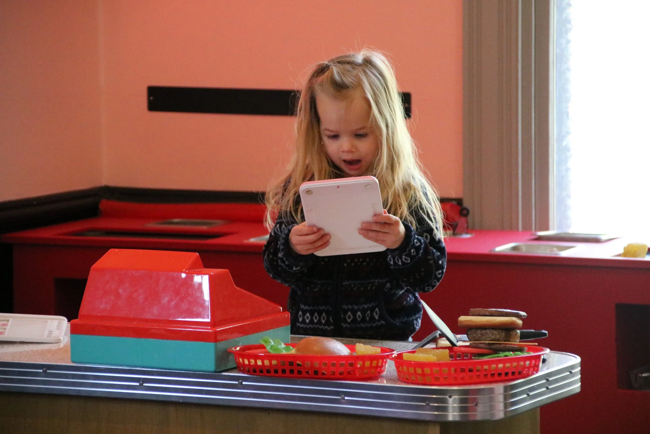 A young girl with long blonde hair looking at a tablet in a play kitchen area with red and blue toy cash register and food items.