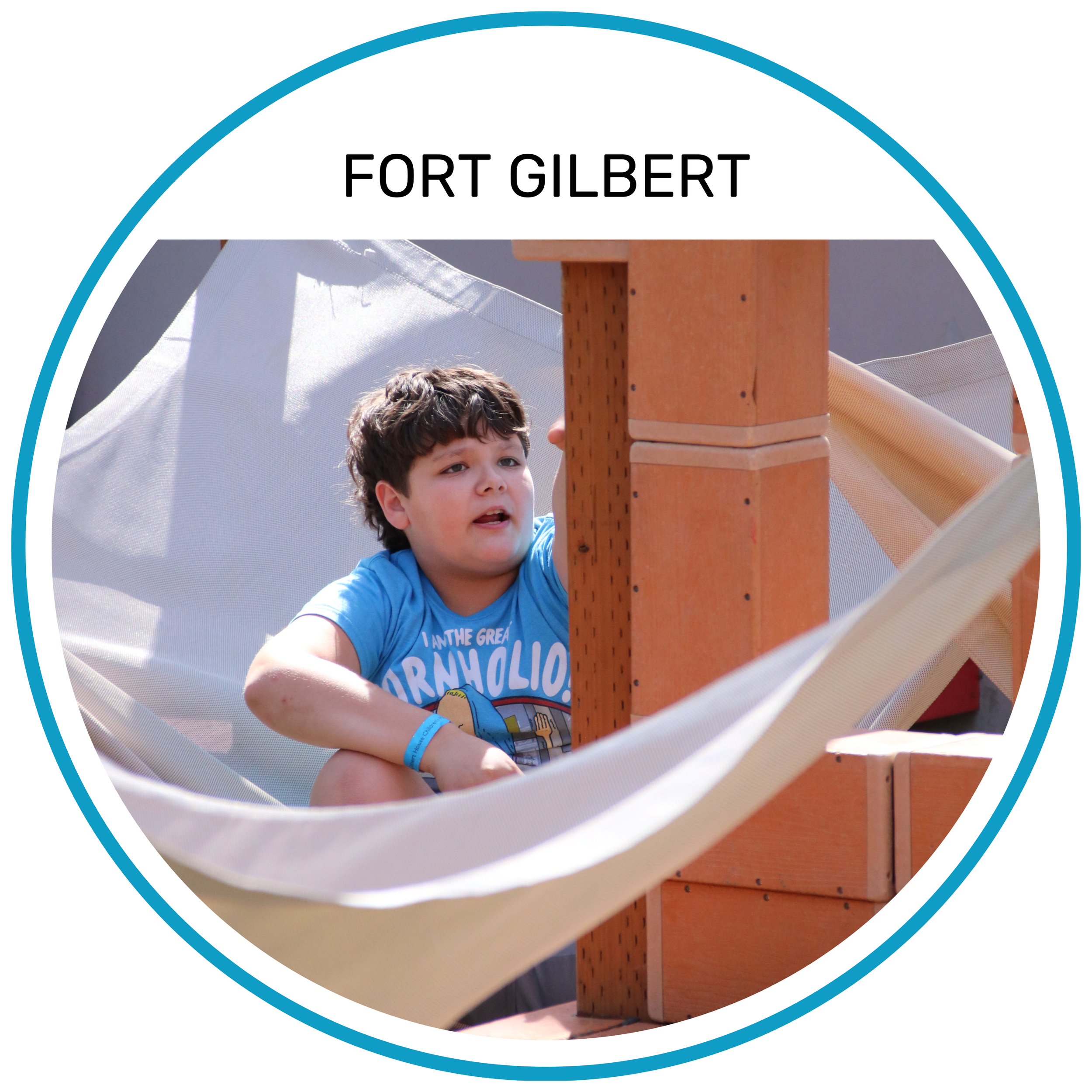 A young boy with curly brown hair wearing a blue shirt sitting on a wooden play structure at Fort Gilbert, looking to the side.
