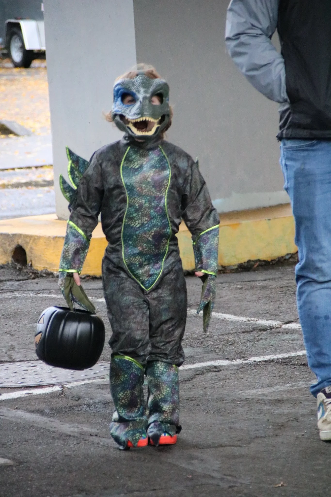 Child dressed in a dinosaur costume with a dinosaur mask, holding a black lunchbox, walking in a parking lot.
