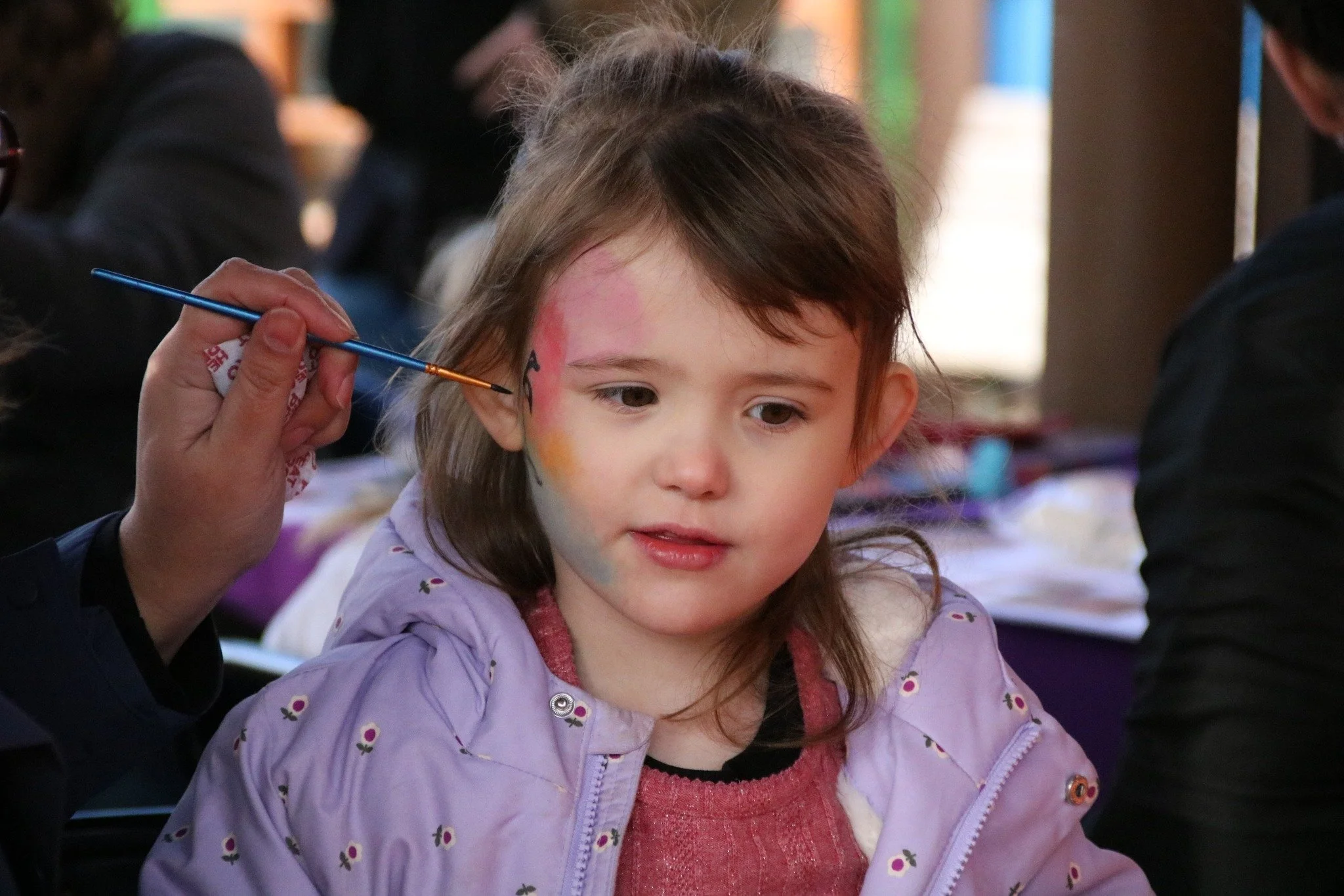 Young girl with face paint being decorated at a community event or festival.