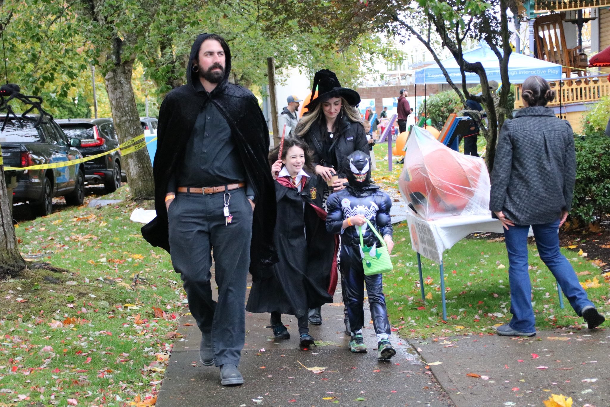 People dressed in Halloween costumes walking outdoors on a cloudy day near a park with trees and fallen leaves.