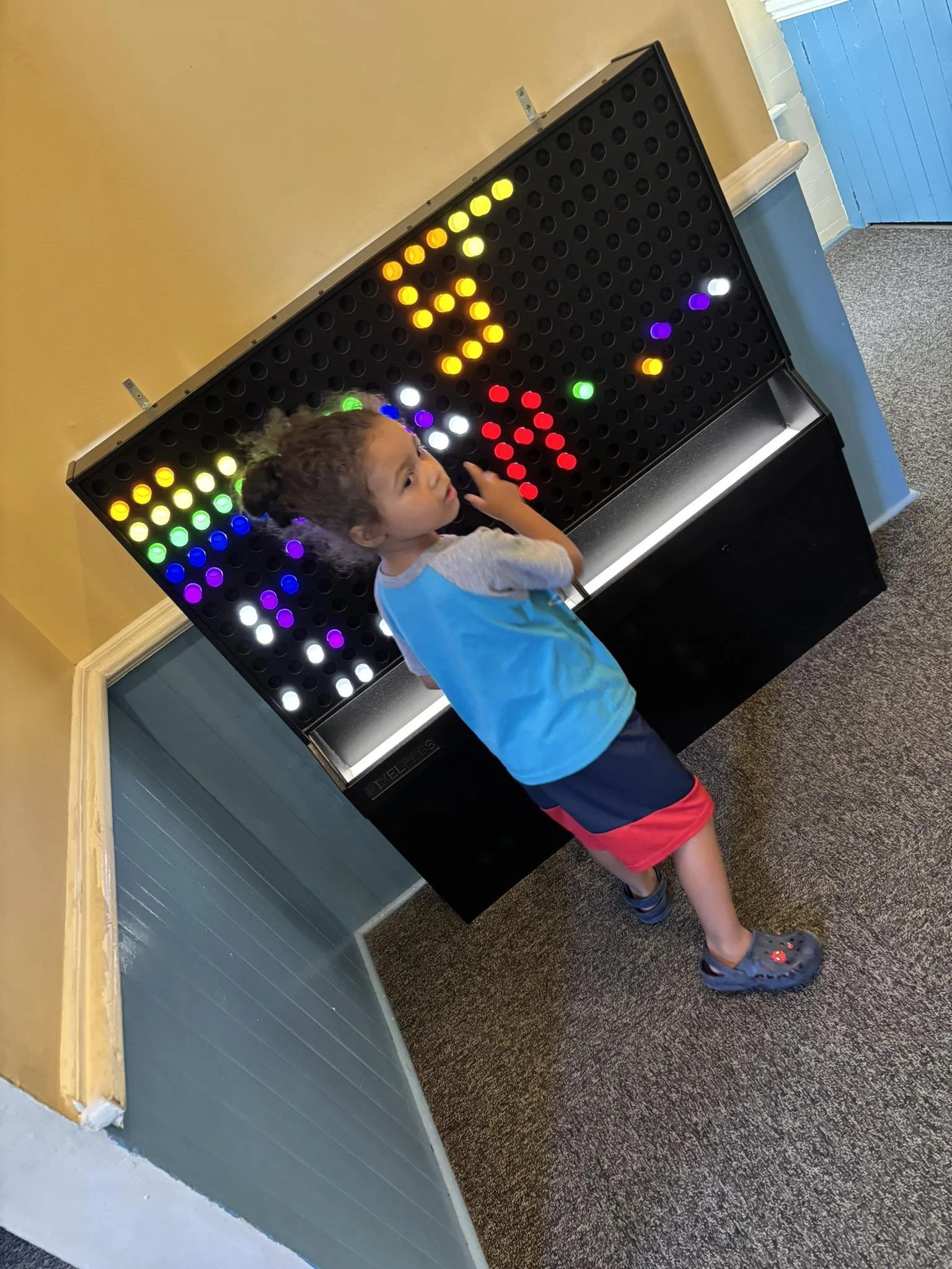 A young child playing a colorful pegboard light game, touching the lights with their right hand, standing on a carpeted floor next to a yellow wall and a blue door.