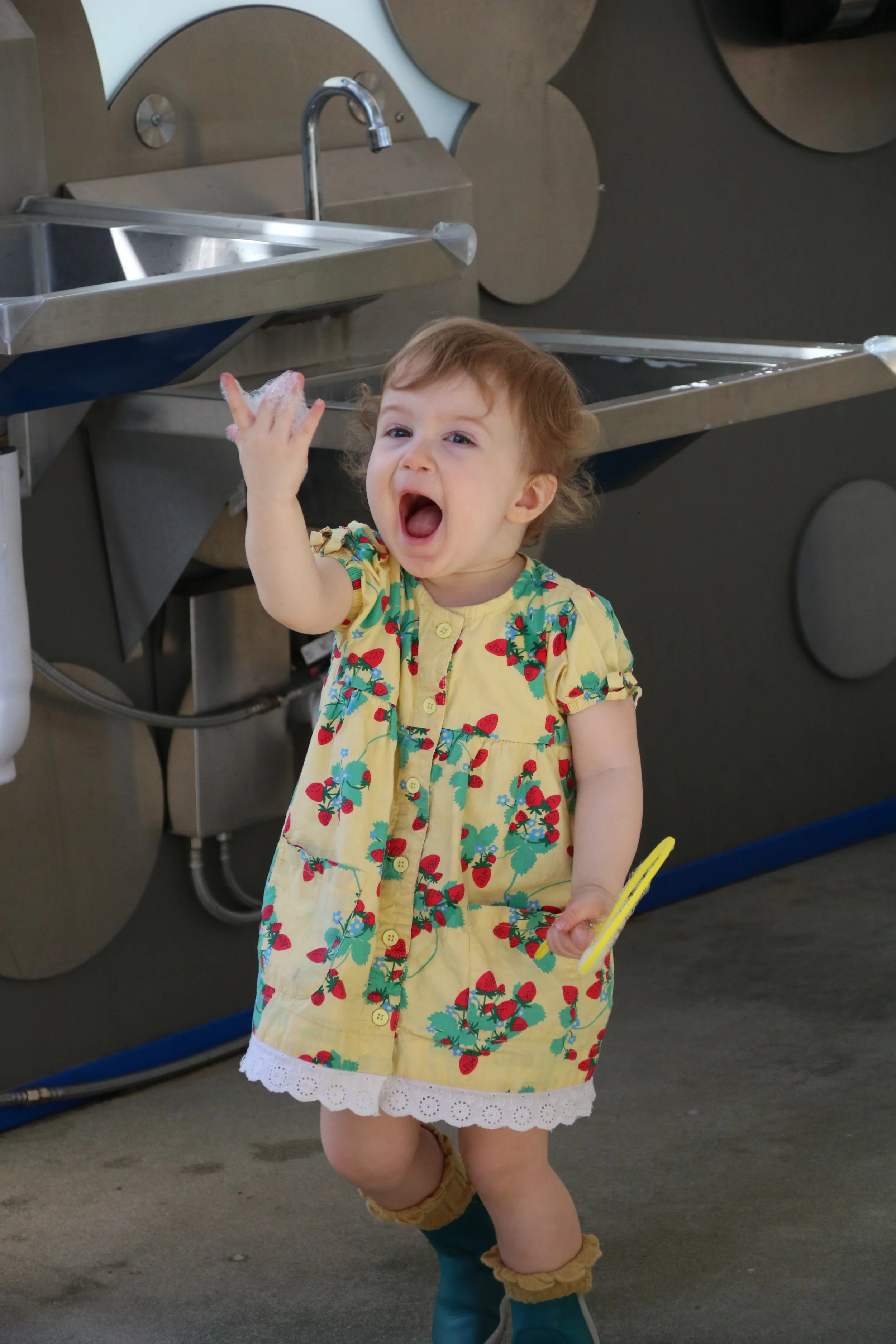 A young girl with curly light brown hair, wearing a yellow dress with a strawberry pattern and lace trim, standing in front of a stainless steel laundry sink, holding a yellow item, and waving with her other hand while smiling.