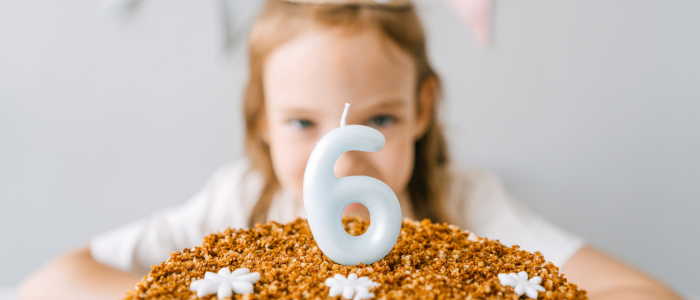 A large, light-blue number "6" candle stands lit in the center of a textured brown birthday cake decorated with delicate white icing flowers. A young girl with light brown hair is visible in the soft-focus background, looking at the cake.