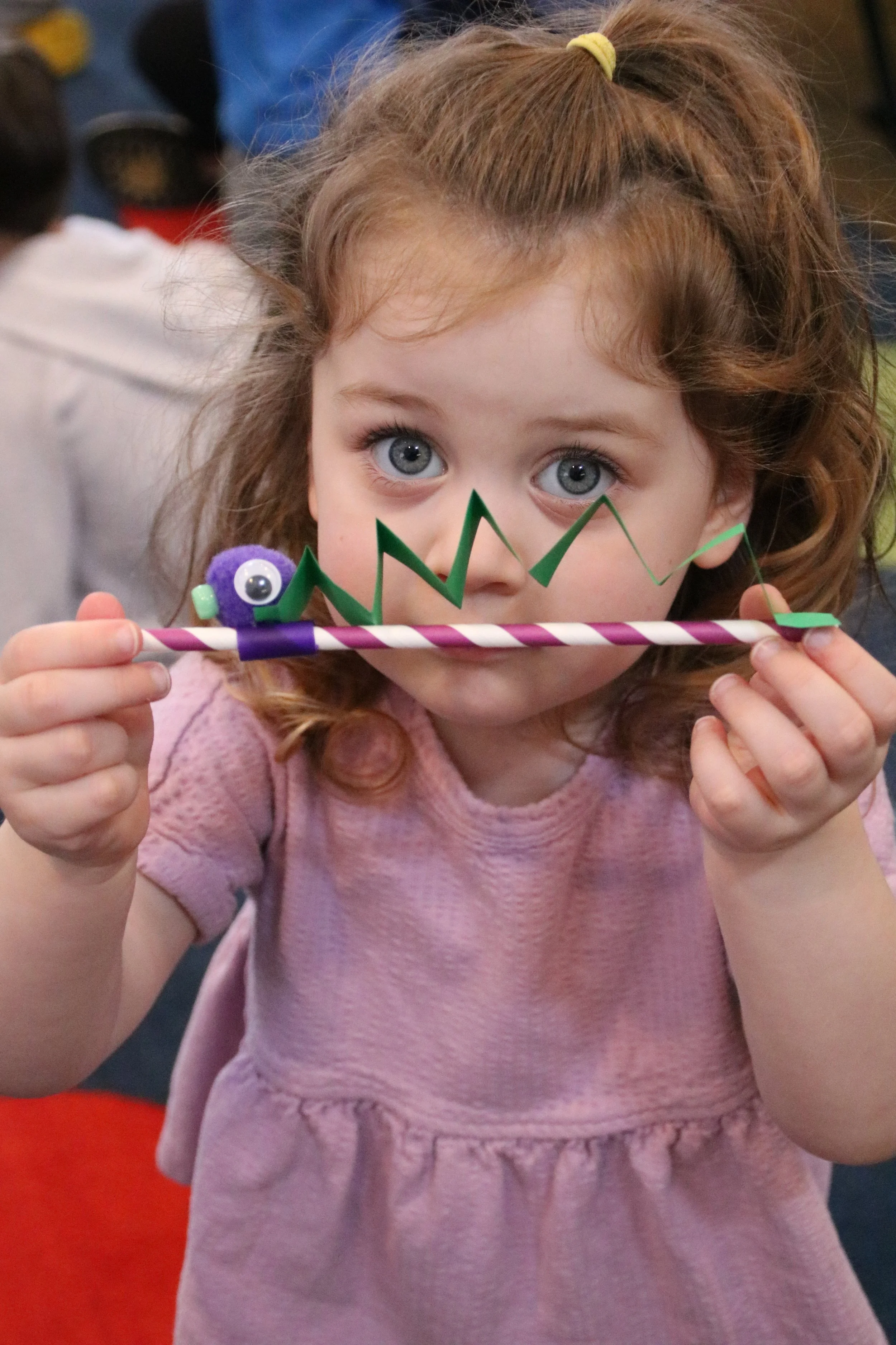 A young girl with curly red hair and blue eyes holding a decorative party horn with a purple and white striped stick and colorful paper decorations, looking directly at the camera.