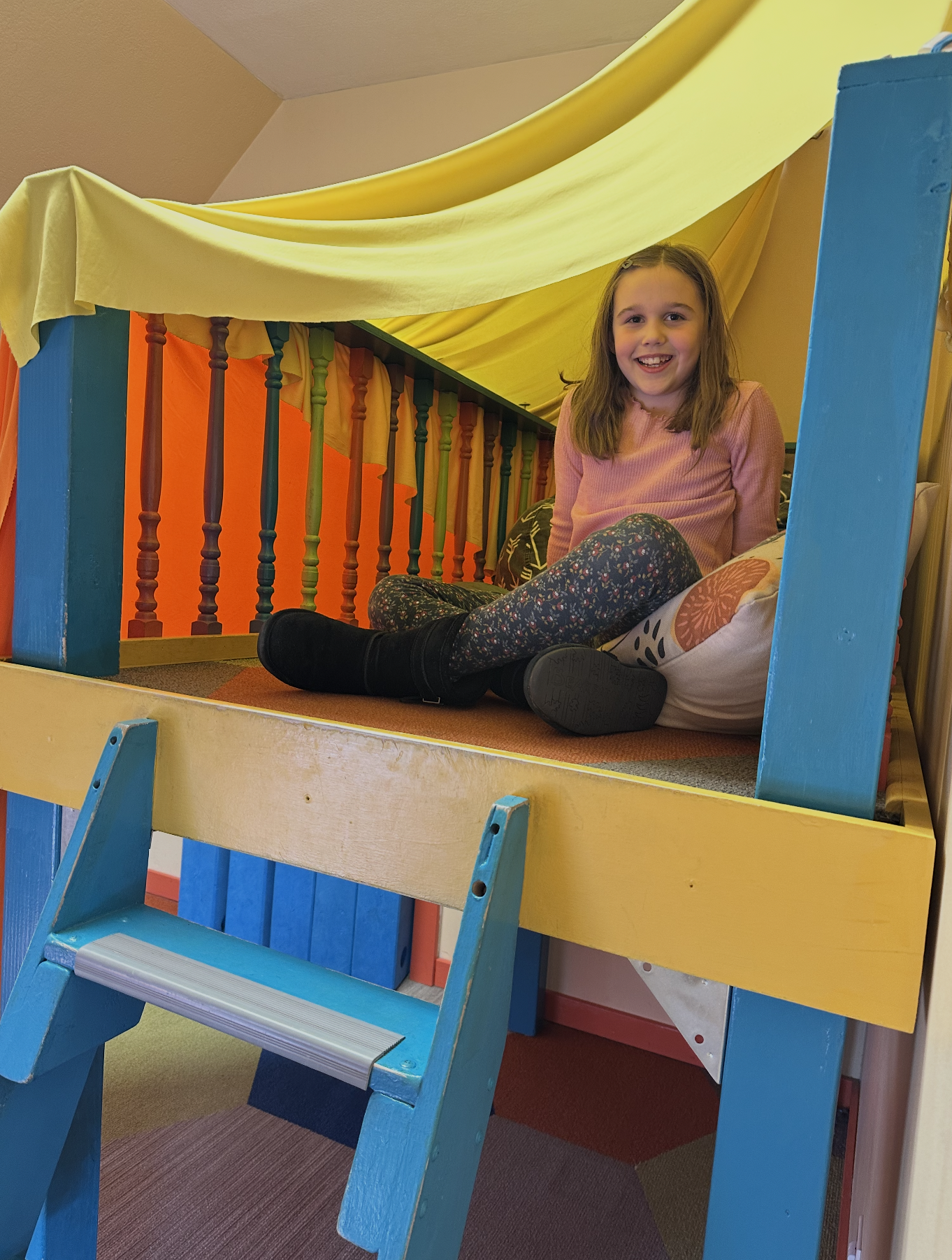 A girl sitting on a colorful indoor play structure with yellow, orange, and blue components, smiling at the camera.