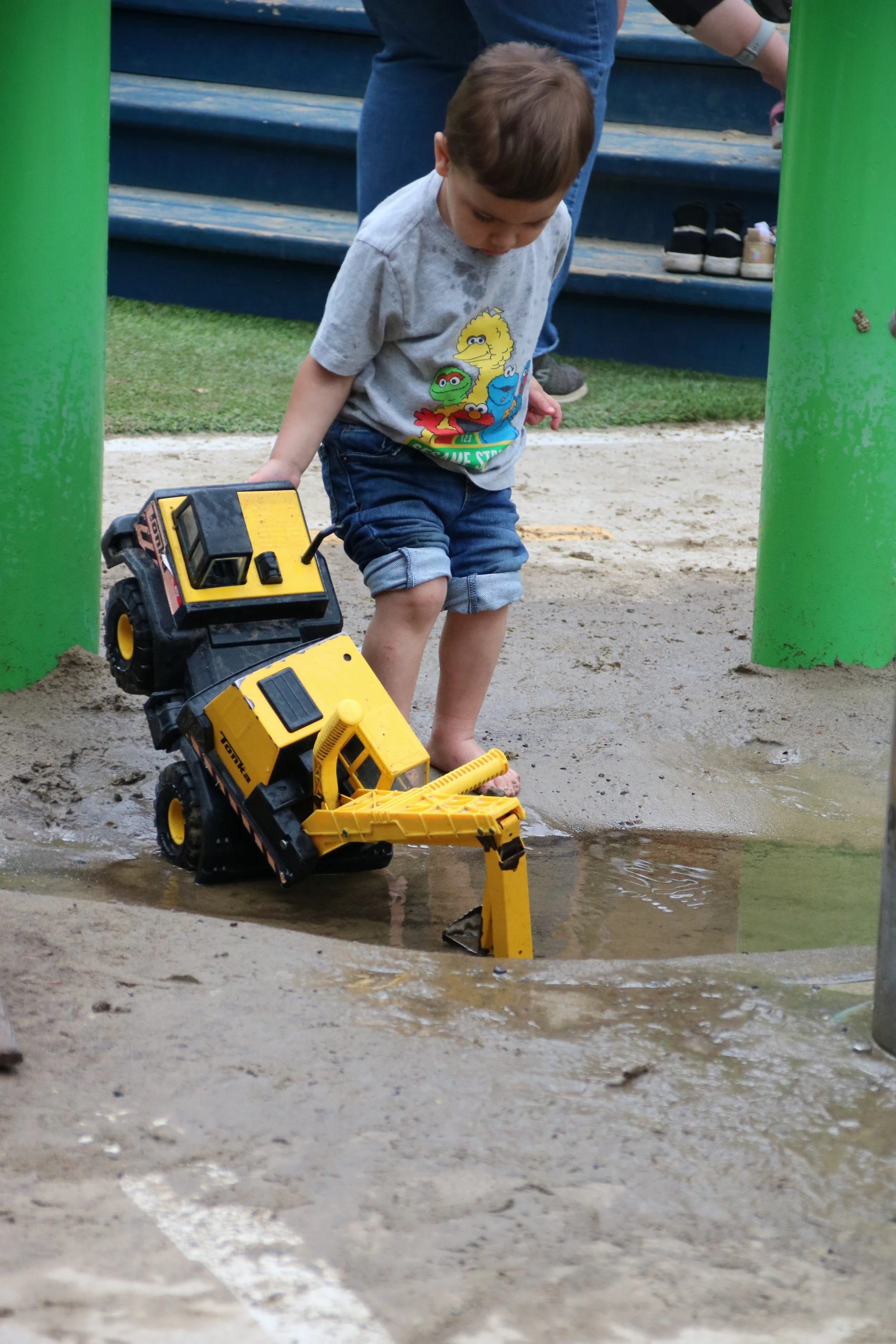 A young boy playing with a yellow toy dump truck in a muddy puddle at a playground.