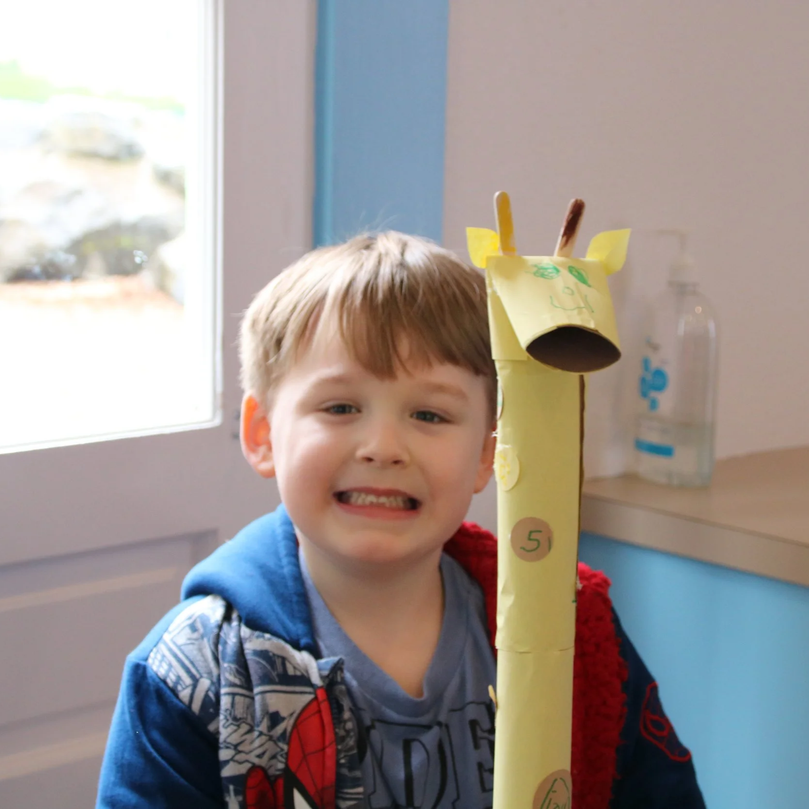 A young boy with red hair smiling next to a homemade paper giraffe craft inside a room with a window and a water bottle on a counter.