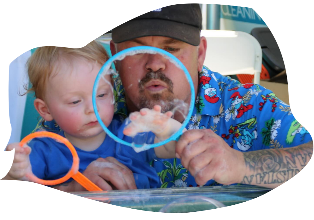 Close-up image of a father and his young son using bright orange wands to create large, shimmering soap bubbles