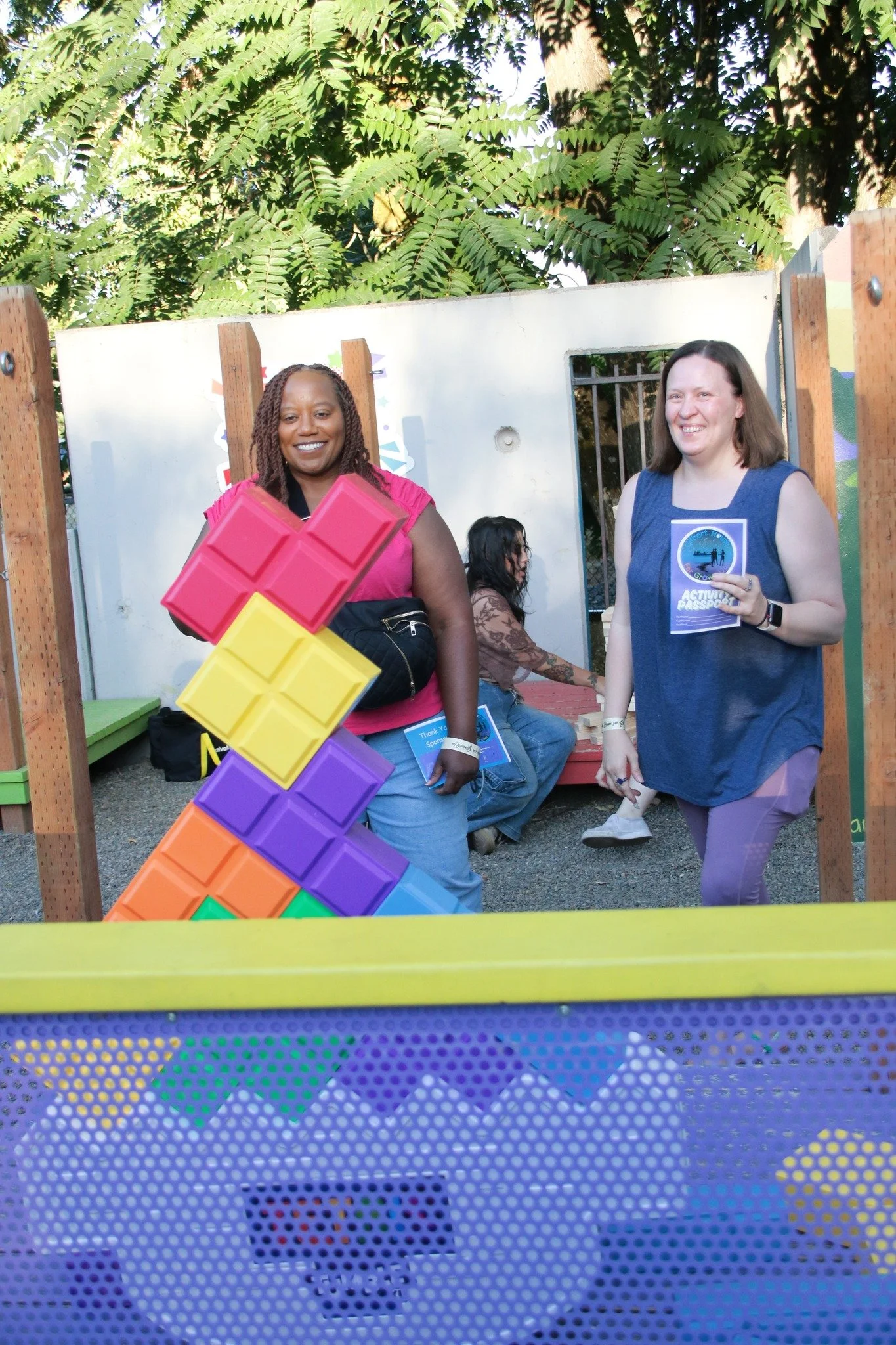 Two women smiling and standing at a booth with large colorful Tetris blocks and a sign for an activity passport, outdoors with trees in the background.