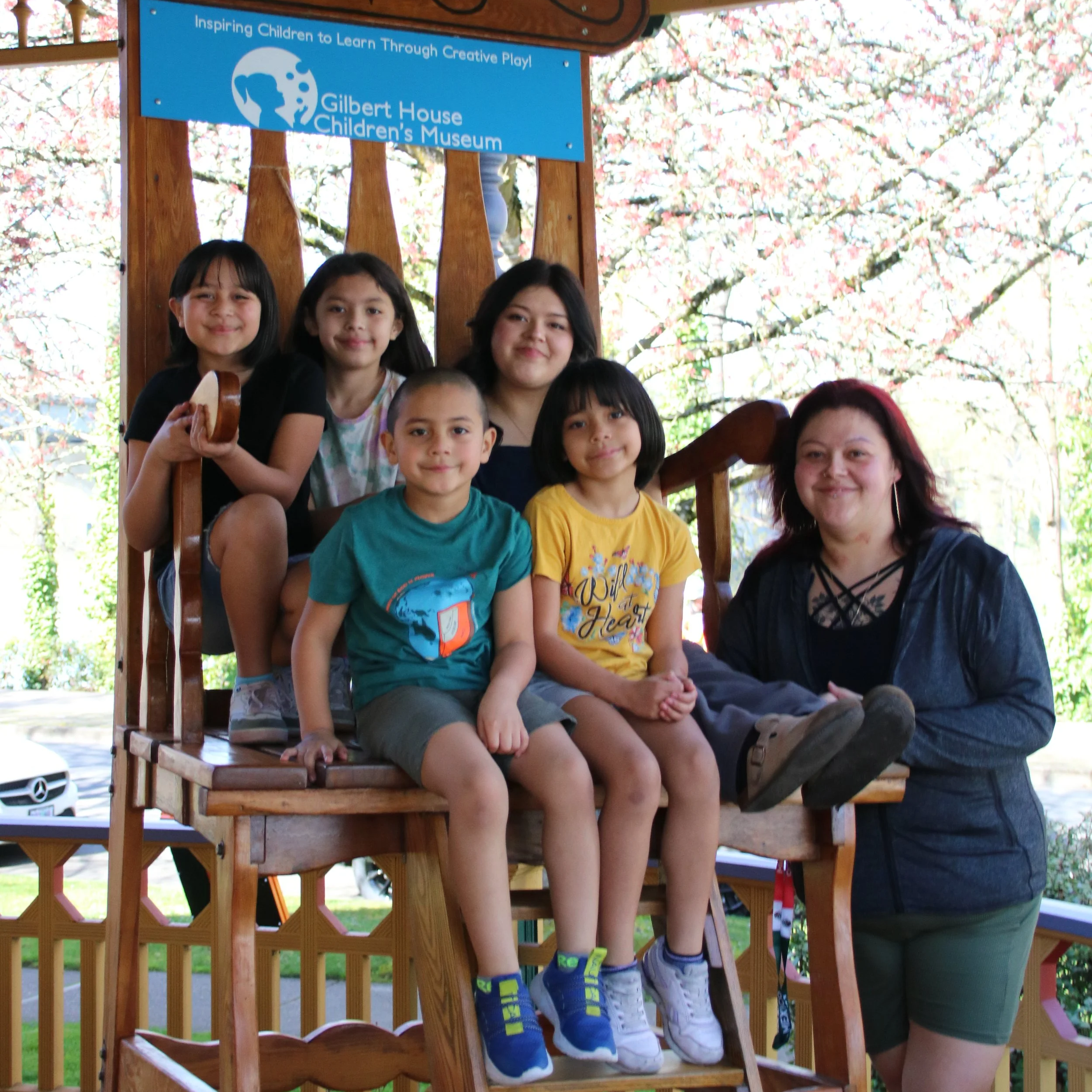 A large, smiling family of six—one adult and five children—posed together on a giant oversized wooden chair outdoors.