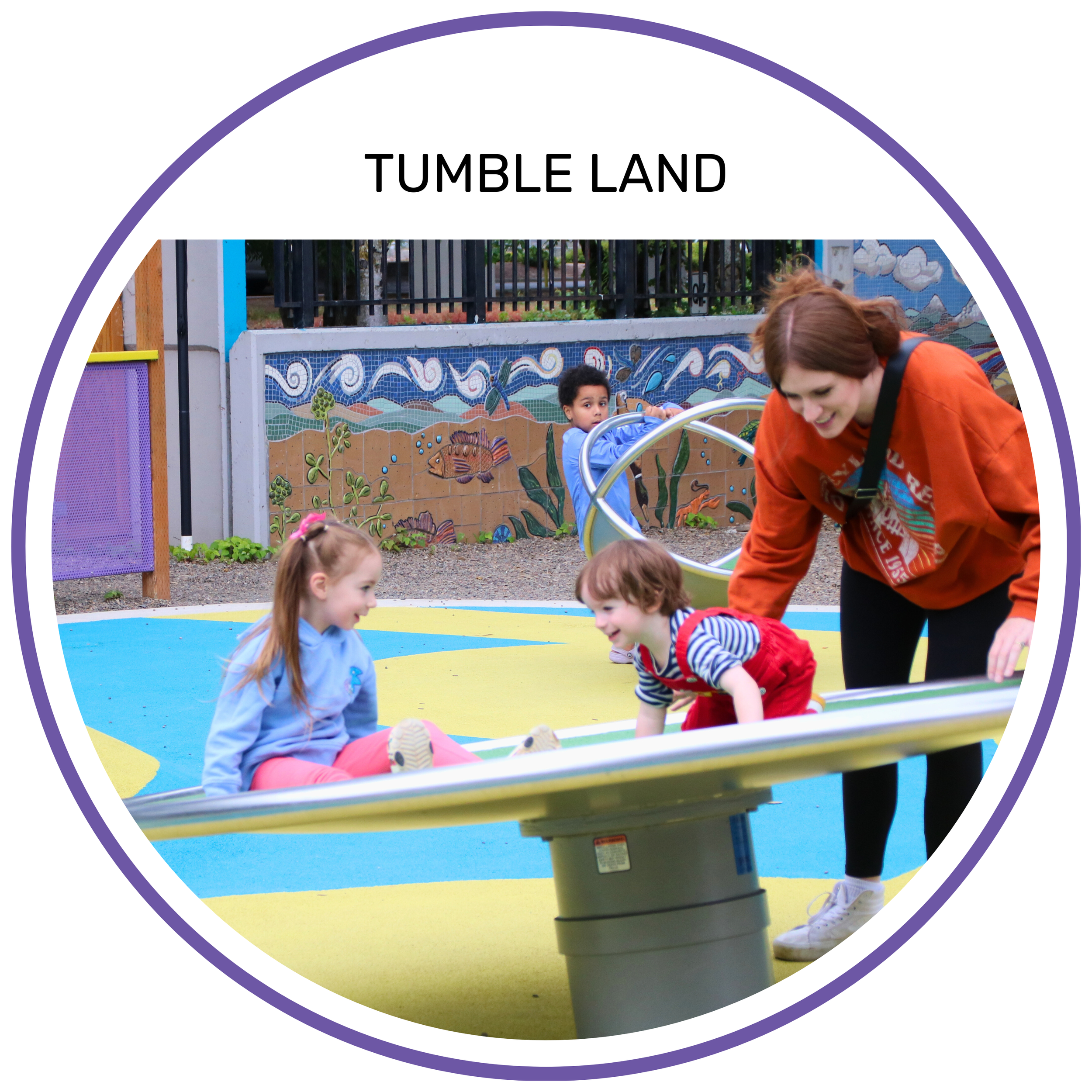 Children and adult playing on a spinning playground equipment labeled 'Tumble Land' in a park.