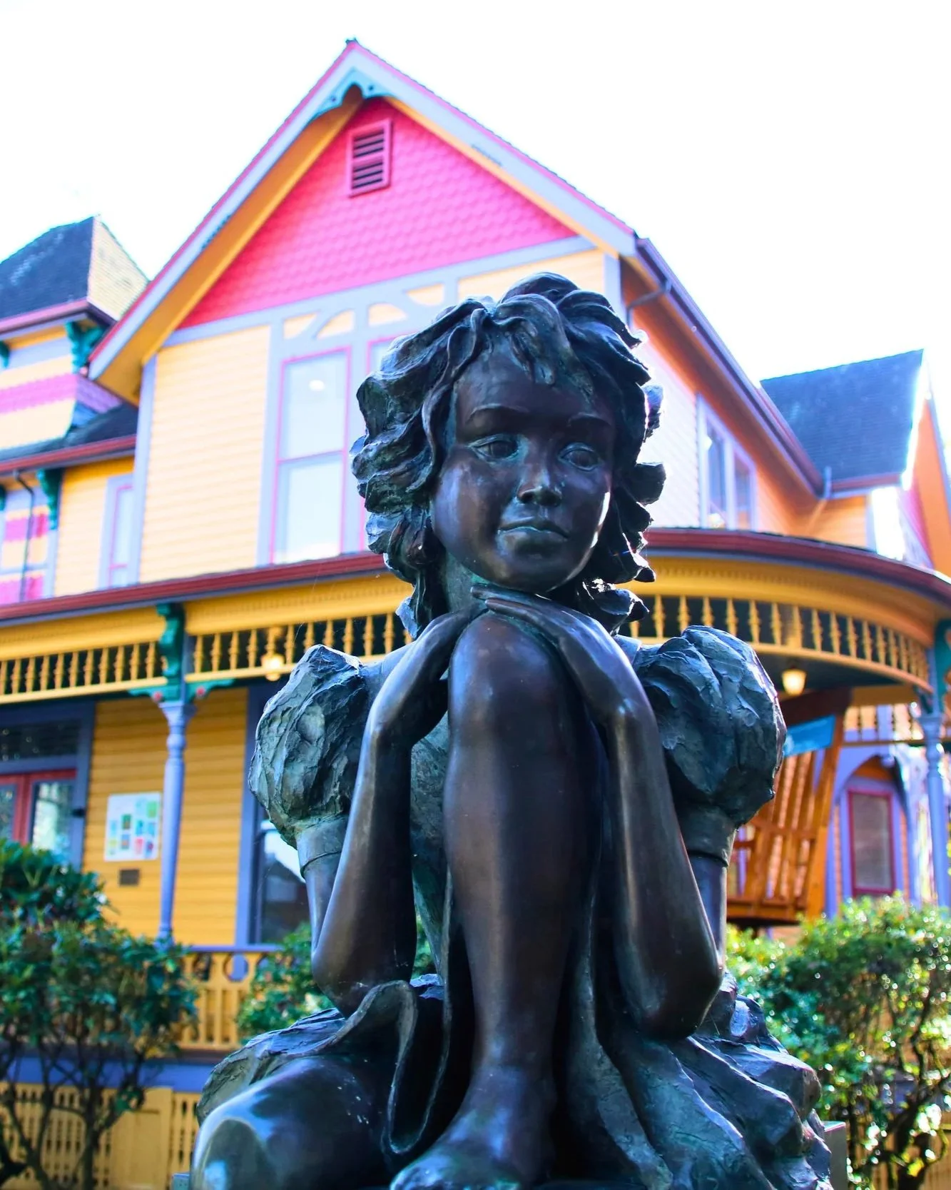 Bronze statue of a girl with curly hair sitting with her hands under her chin, in front of a colorful house with yellow walls and pink trim.