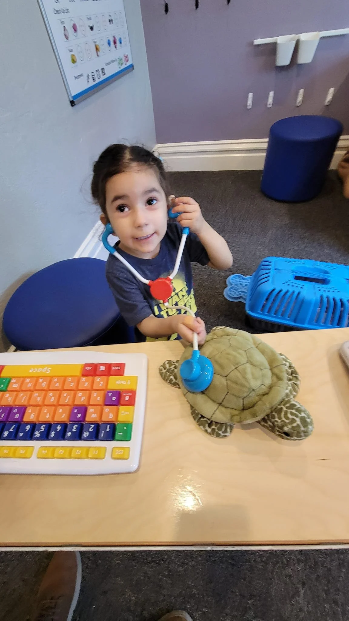 A young girl in a dark shirt is sitting at a table, pretending to listen to a turtle plush toy with a toy stethoscope, in a room with educational decorations on the wall.