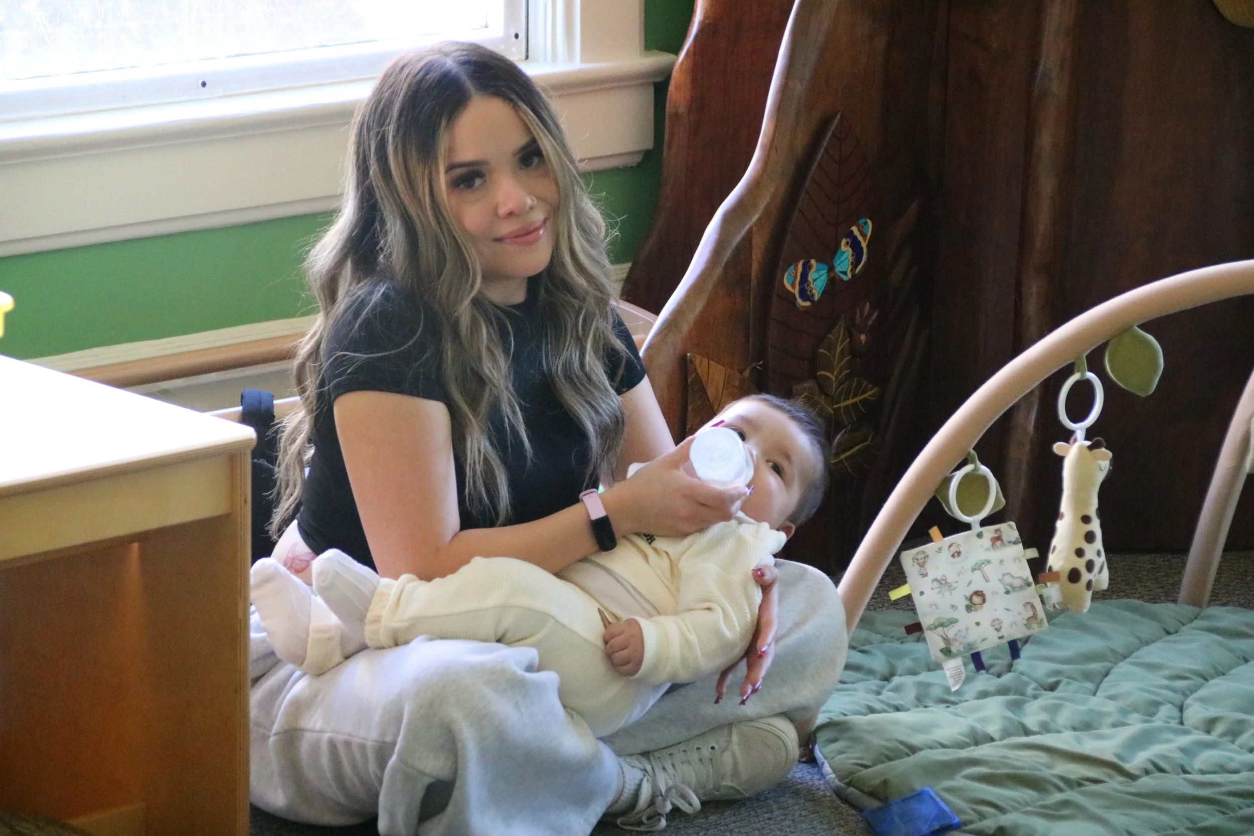 A woman with long wavy hair sitting on the floor, feeding a young child with a bottle. The child is lying on her lap. The background shows a green wall, a window, and a wooden crib with a giraffe toy and a hanging book. The woman is smiling and wearing a black T-shirt.