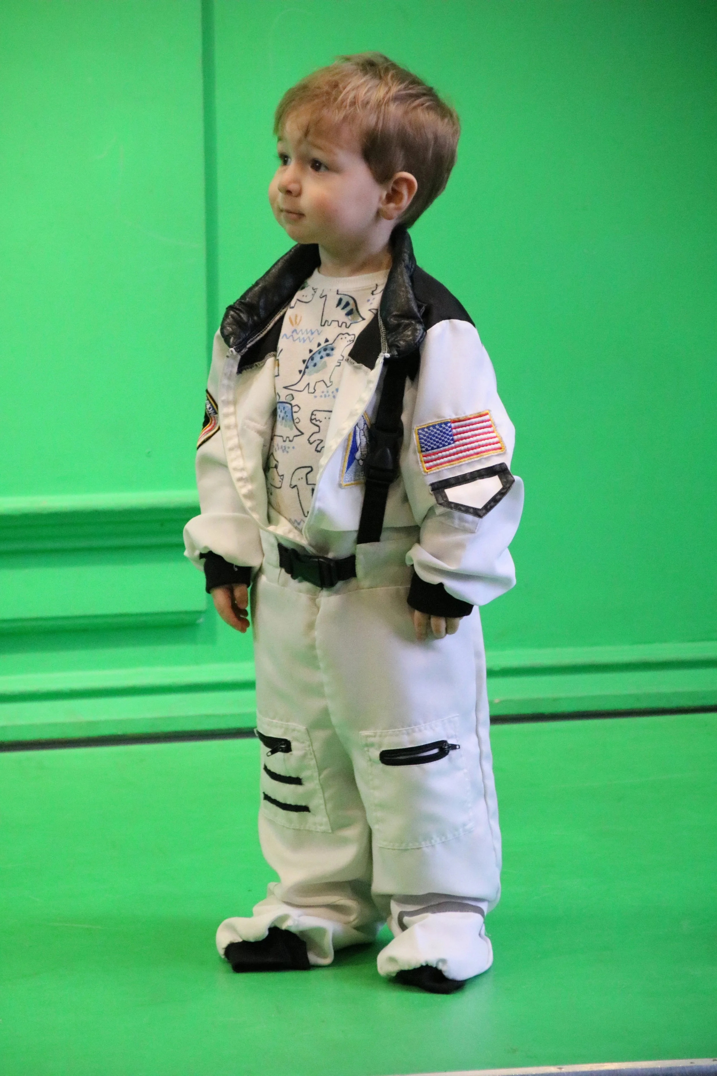 Young boy dressed in a white astronaut costume with patches, standing against a green background with a thoughtful expression.