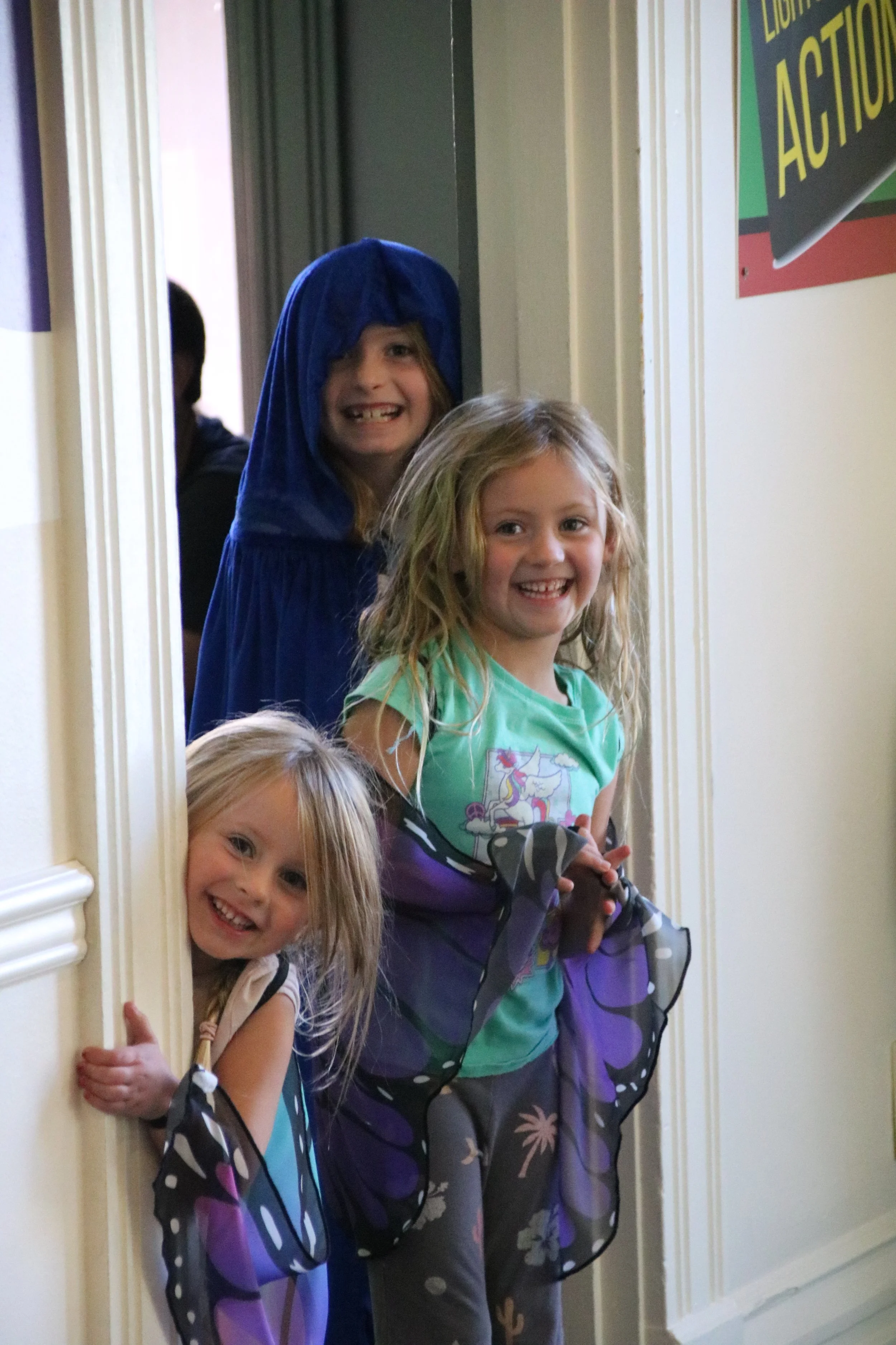 Three young girls peeking around a door frame, smiling and laughing, with one girl dressed in a blue costume with a hood and two girls in casual costumes with fairy wings.