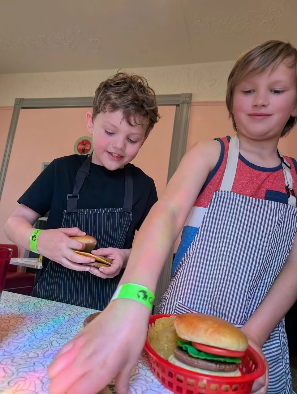 Two young boys wearing striped aprons preparing cheeseburgers at a table.