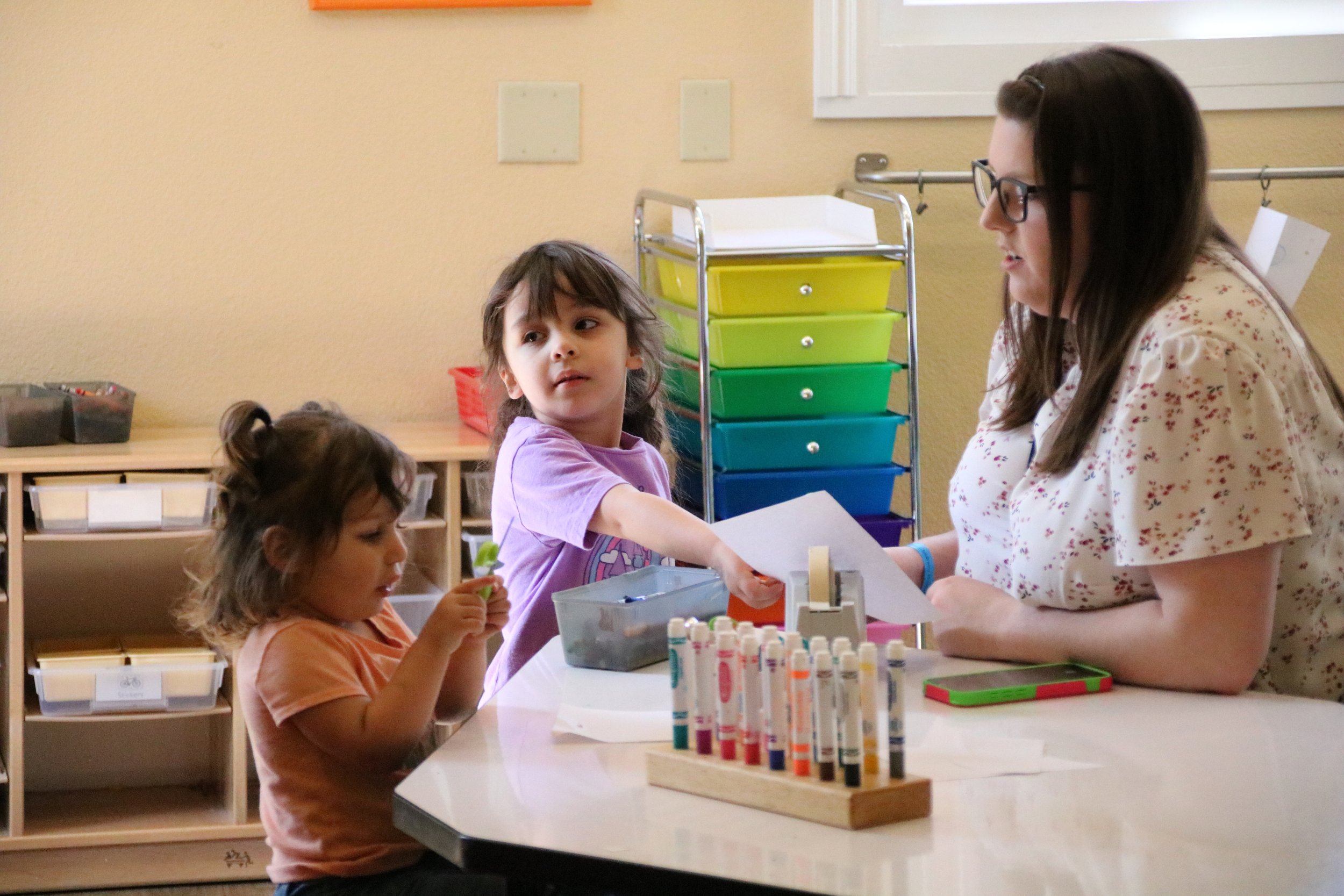 A teacher interacts with two young girls in a classroom. One girl with dark hair and a purple shirt reaches out, while the other girl with curly hair in a peach shirt looks at a small object in her hands. The classroom has colorful storage drawers and art supplies.