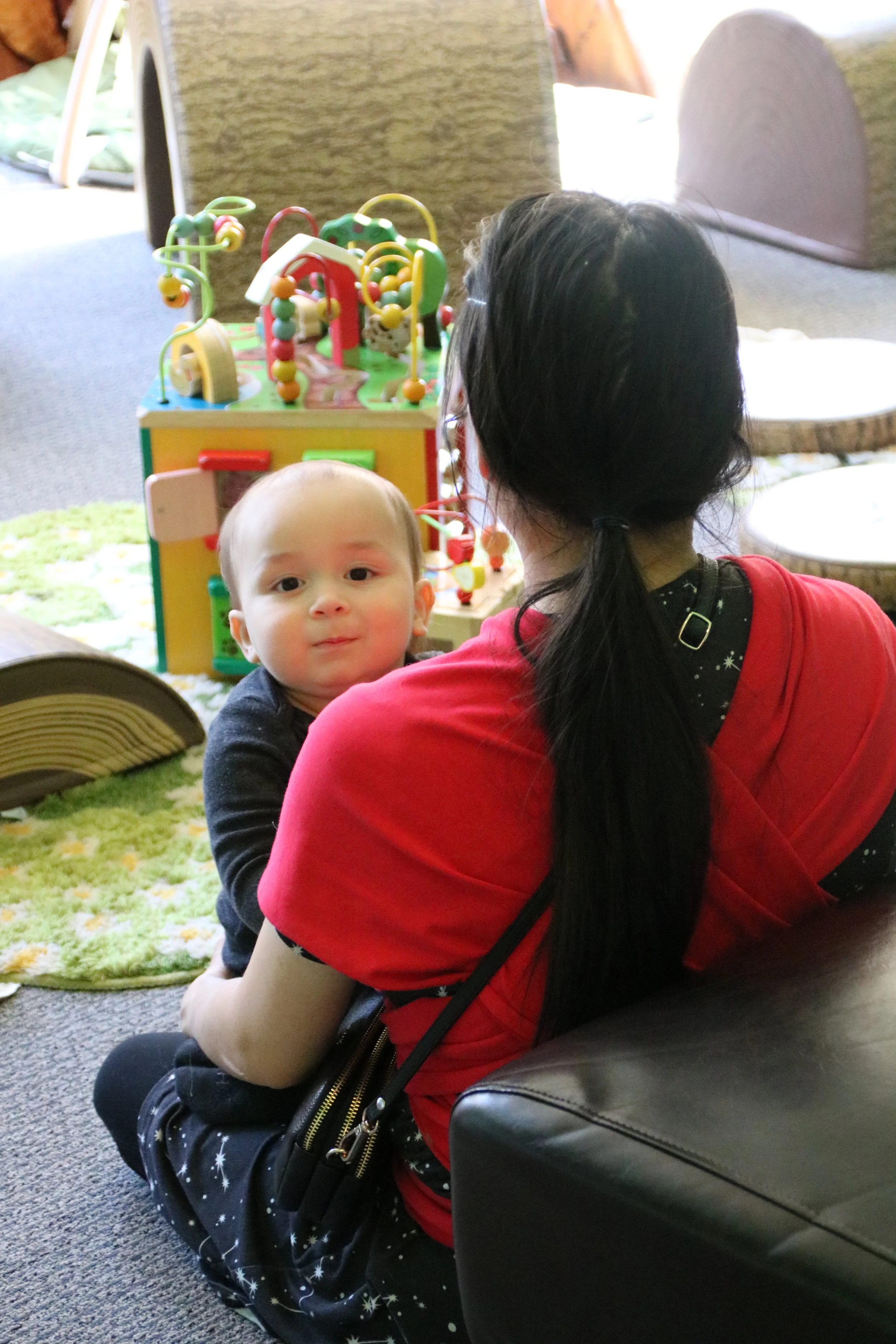 A woman with long black hair in a ponytail, wearing a red shirt, is sitting on a brown sofa holding a smiling baby with a bald head wearing a black outfit. They are in a play area with a colorful bead maze toy and a circular green rug. In the background, there are some furniture pieces and a window.