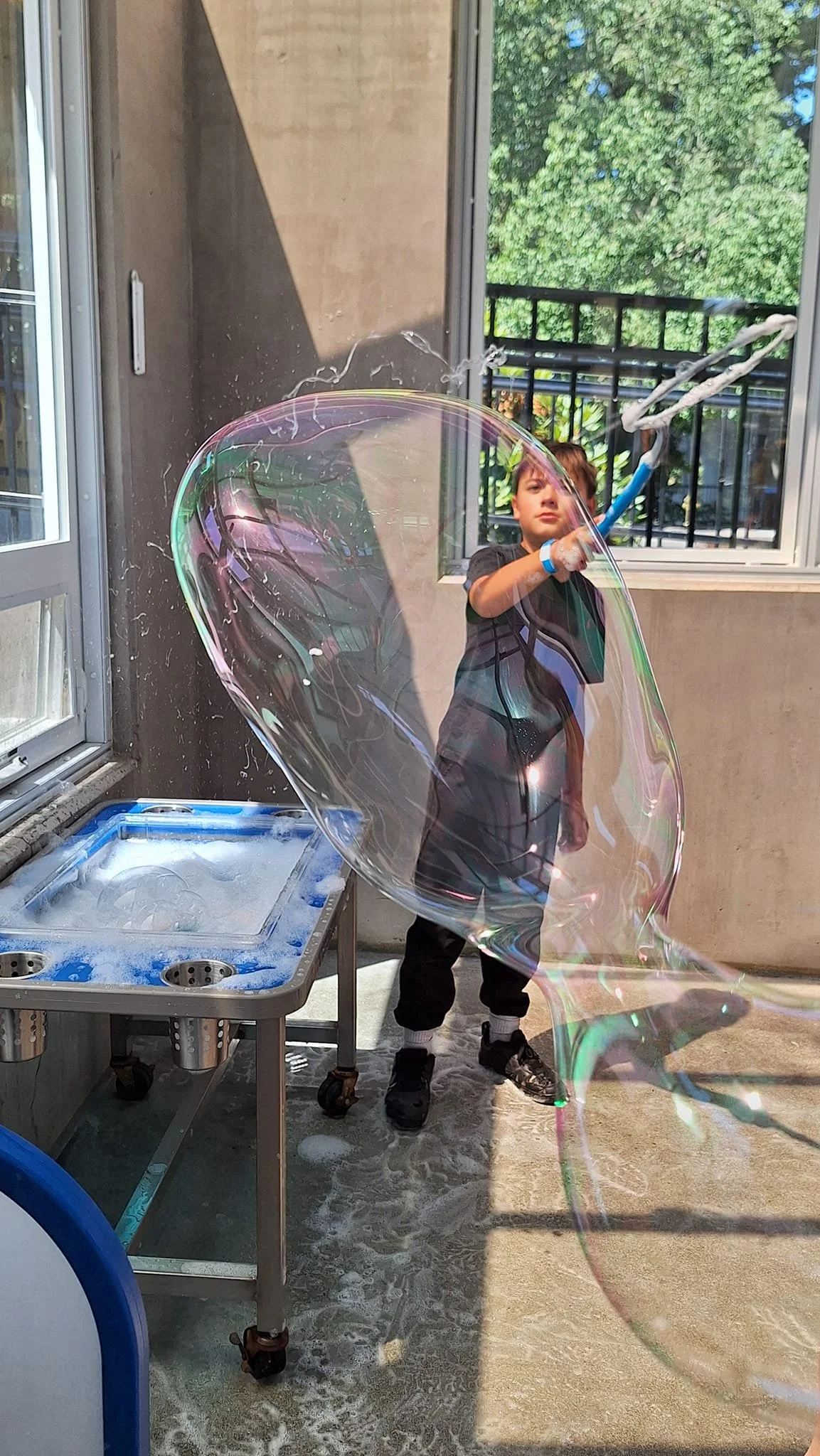 A young boy blowing a large soap bubble with a bubble wand inside a bright room with windows and a view of green trees outside.