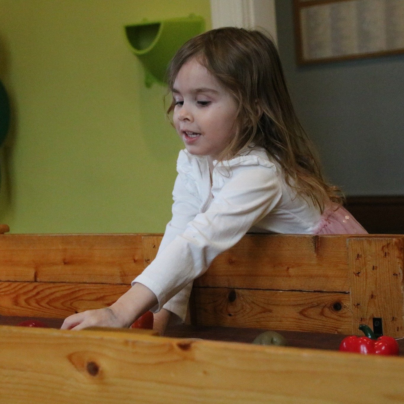 A young girl with light brown hair reaching to her shoulders is playing a wooden arcade game with plastic vegetables, including red peppers, in a room with green and gray walls.