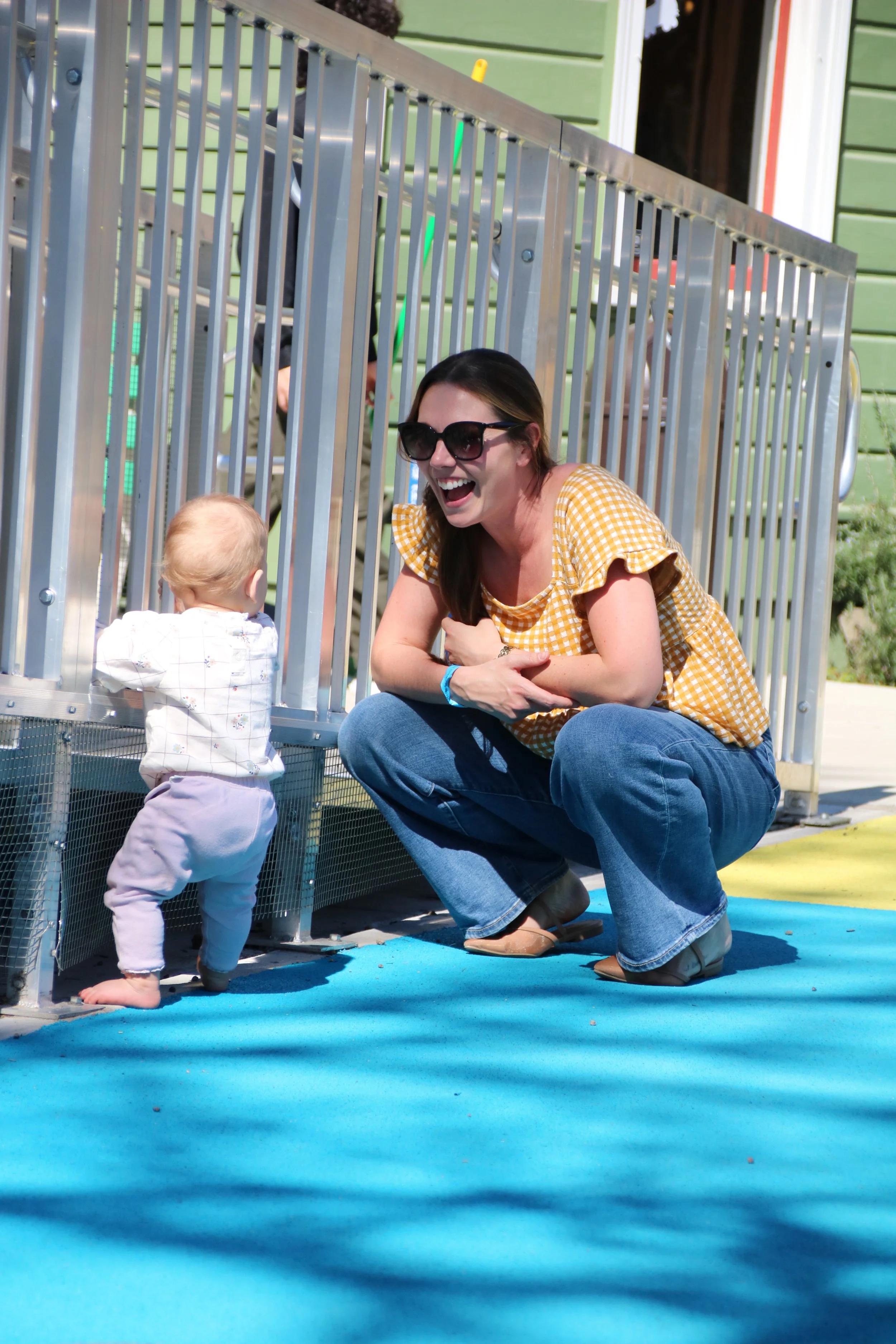 A woman crouching down and smiling at a small child near a metal fence at an outdoor play area.