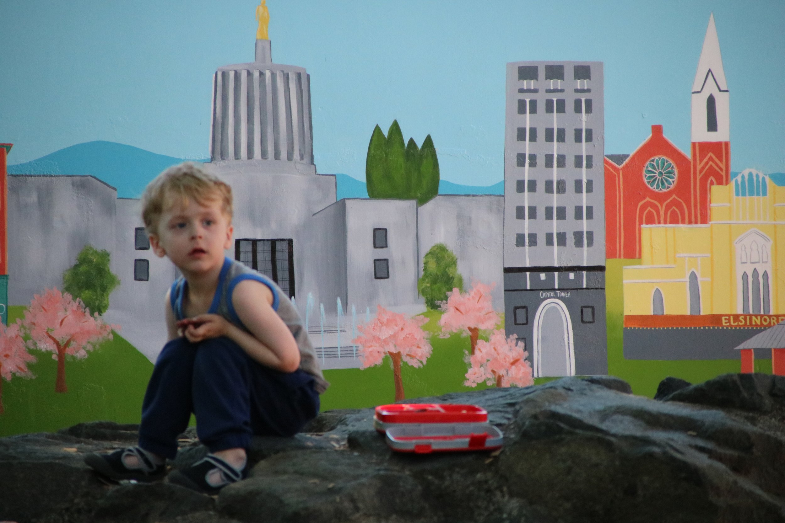 A young boy with curly blonde hair, wearing a gray sleeveless shirt and dark pants, is crouching on a rocky surface in front of a colorful city mural. The mural depicts buildings, trees, and mountains, including a gray modern building, a red church, and pink blossoming trees.