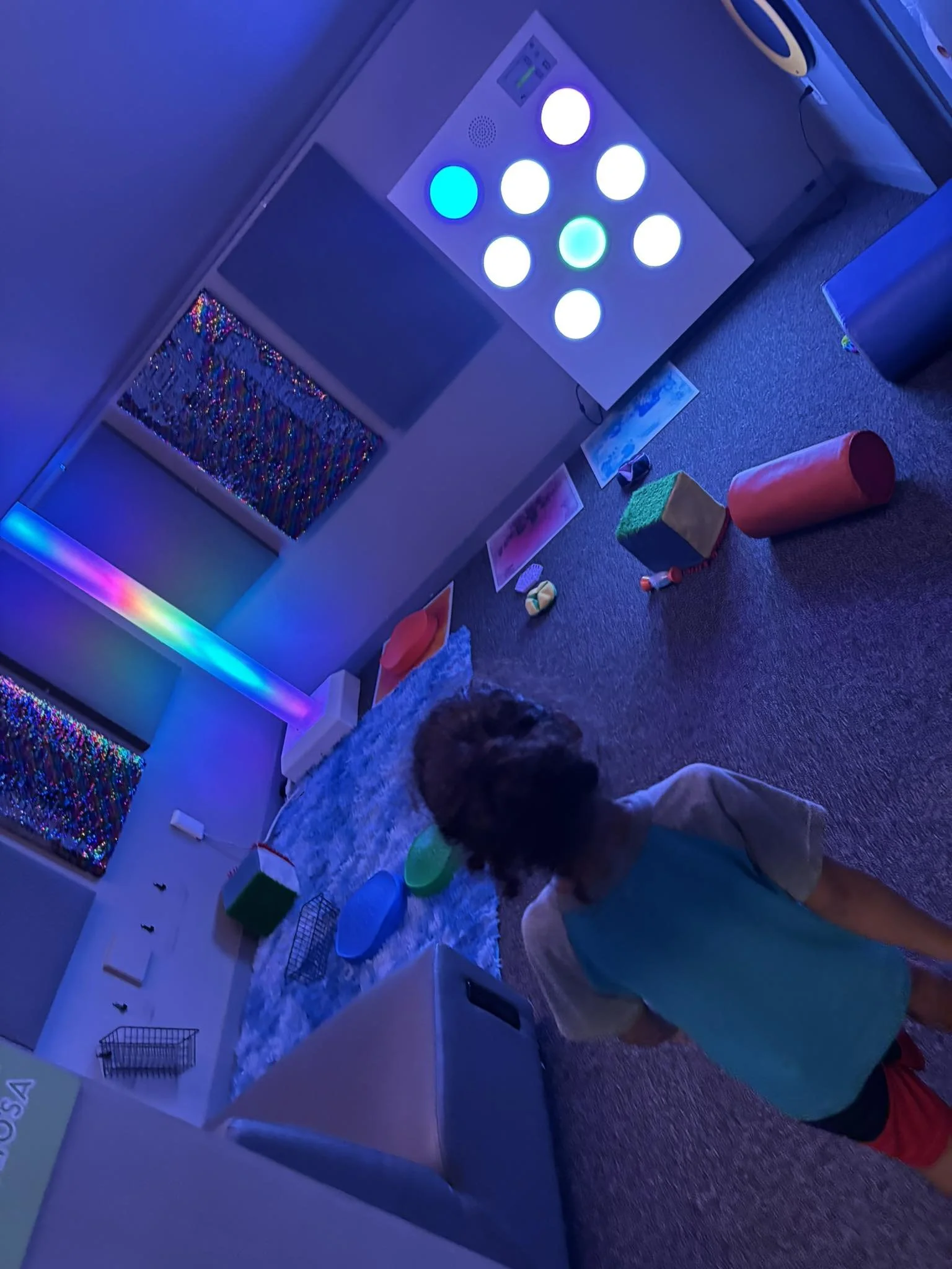 Child standing in a dark room with colorful LED lights and various toys and objects around.