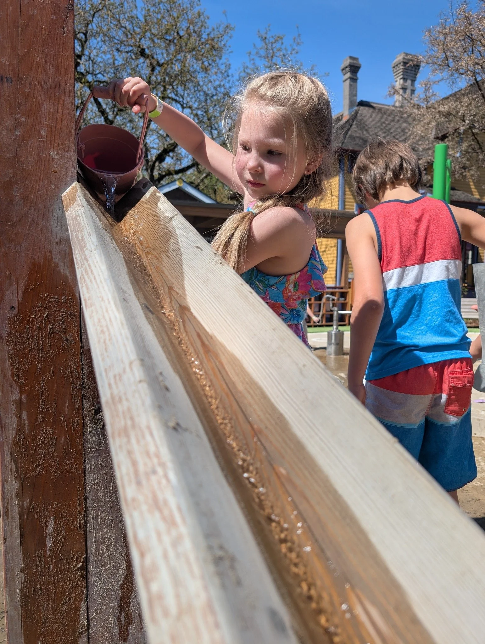A young girl pouring water into a wooden water slide at a playground while a boy stands nearby. It is a sunny day with trees in the background.