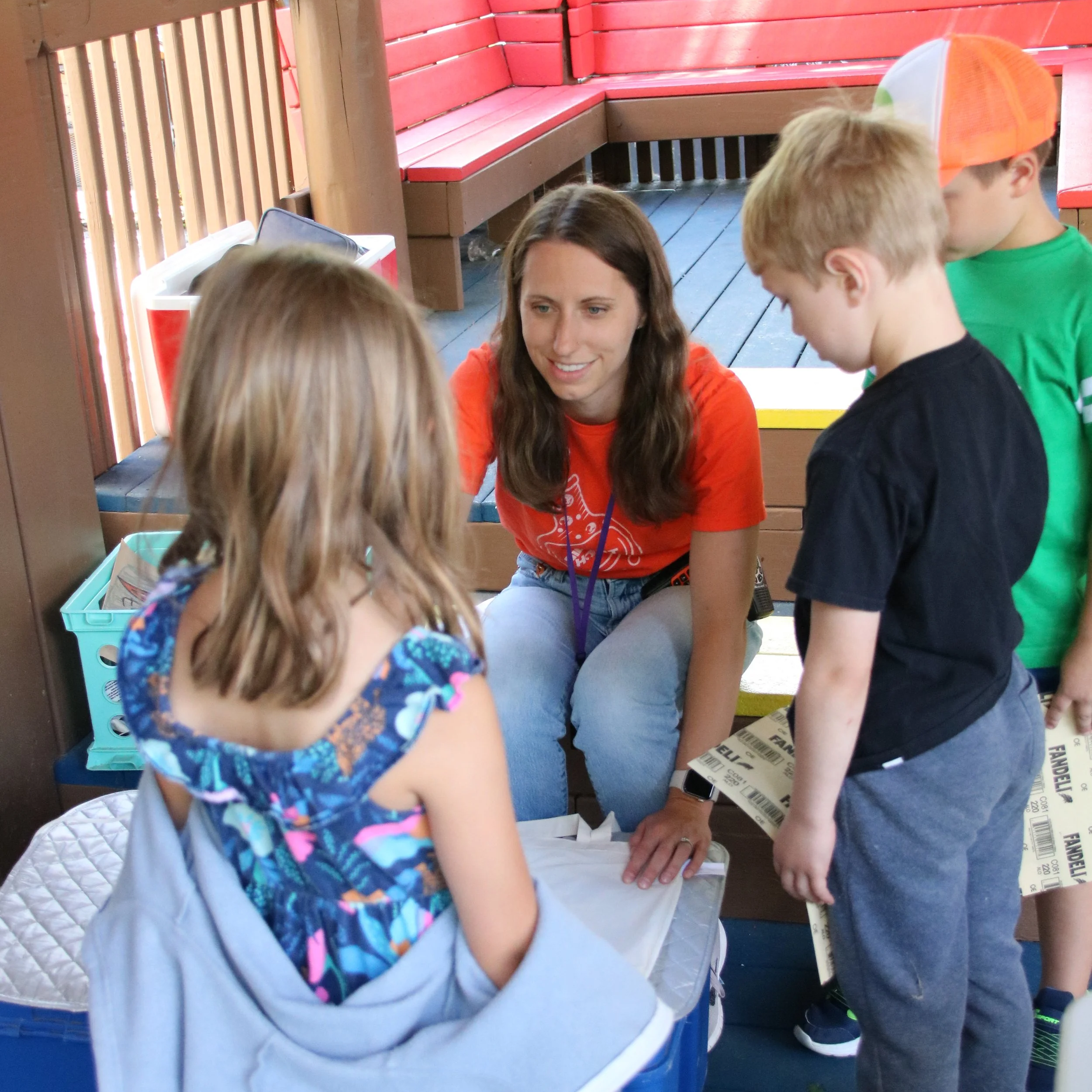 A woman talking to three children at an outdoor event, with benches and colorful wooden structures in the background.