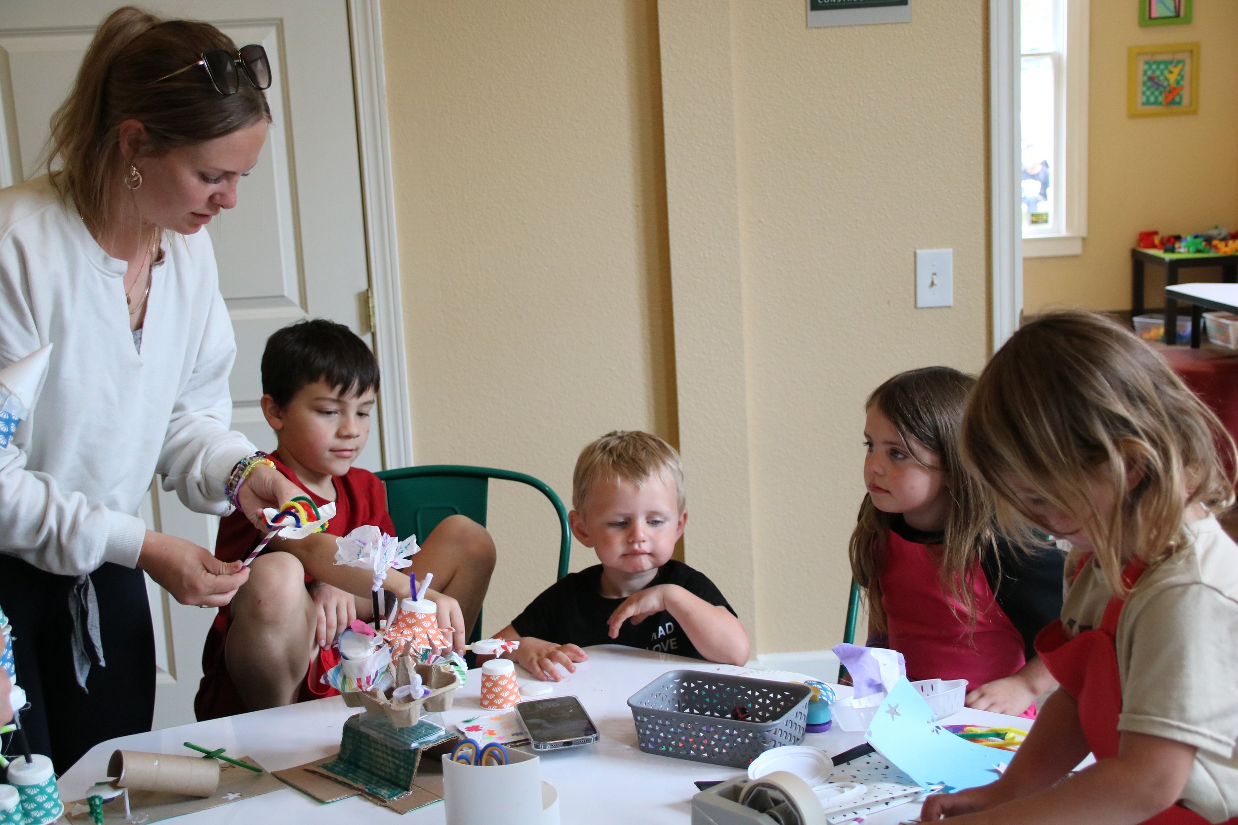 A woman and four children gathered around a table with craft supplies, working on a craft project during a children’s party.
