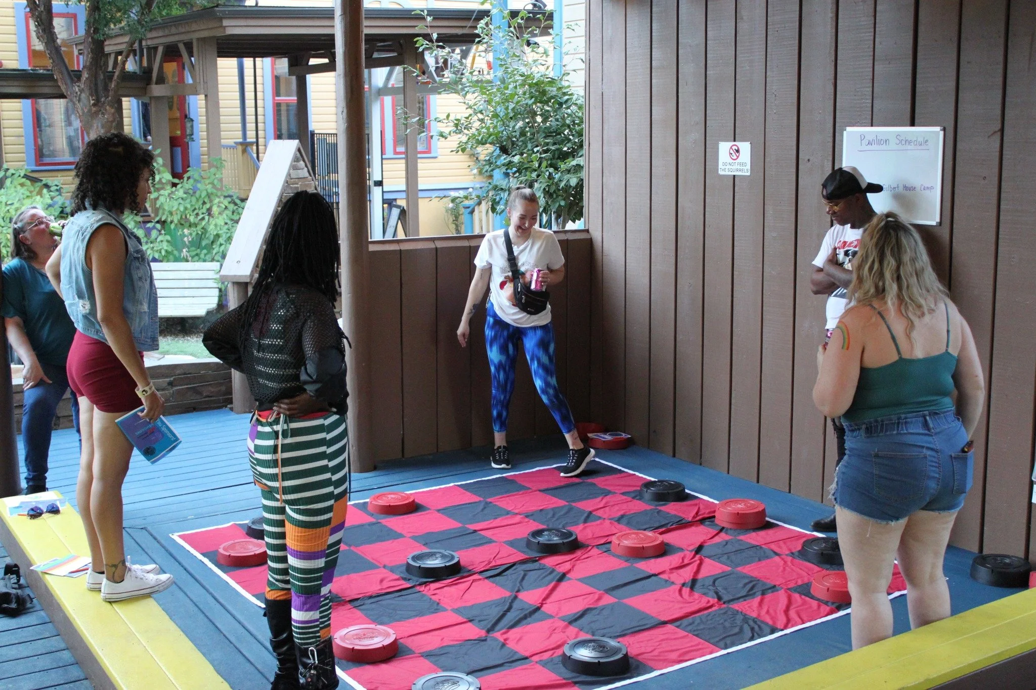 Group of people playing a game of Twister on a red and black mat outdoors on a porch, with a woman in white and blue leggings in the middle.