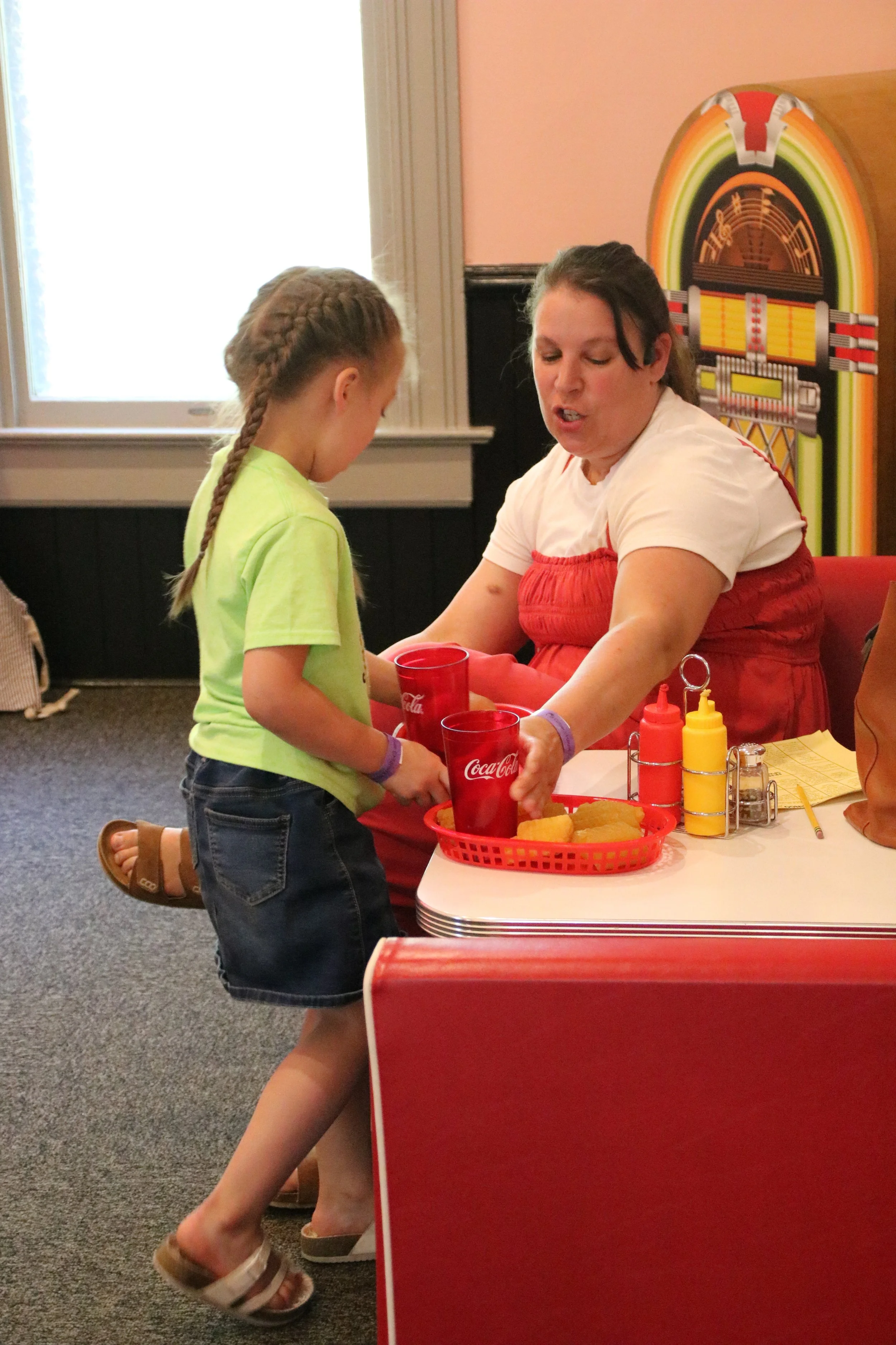 A young girl with braided hair, wearing a neon green t-shirt and denim shorts, is standing at a counter, being served food by a woman at a retro-style diner. The woman is wearing a white t-shirt with a red apron and is handing over two red Coca-Cola cups.