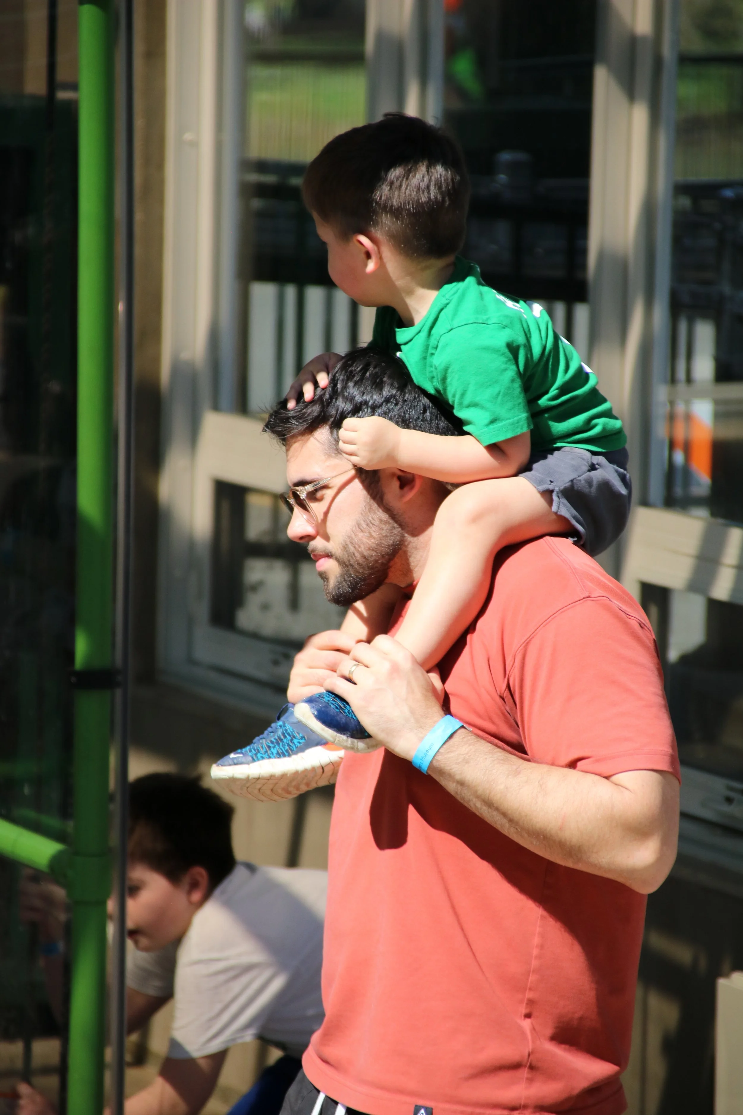 A man with dark hair, glasses, and a beard, wearing a salmon-colored t-shirt, is carrying a young boy on his shoulders. The boy has short dark hair, is wearing a bright green t-shirt and gray shorts, and is holding the man's head with one hand. They are outdoors in a sunny area, near a building with large windows.