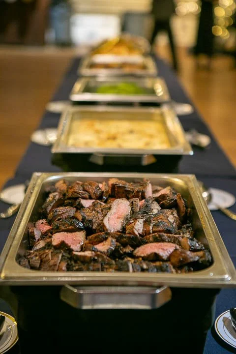 Chafing dishes with various hot foods on a buffet table, including sliced beef in the foreground.