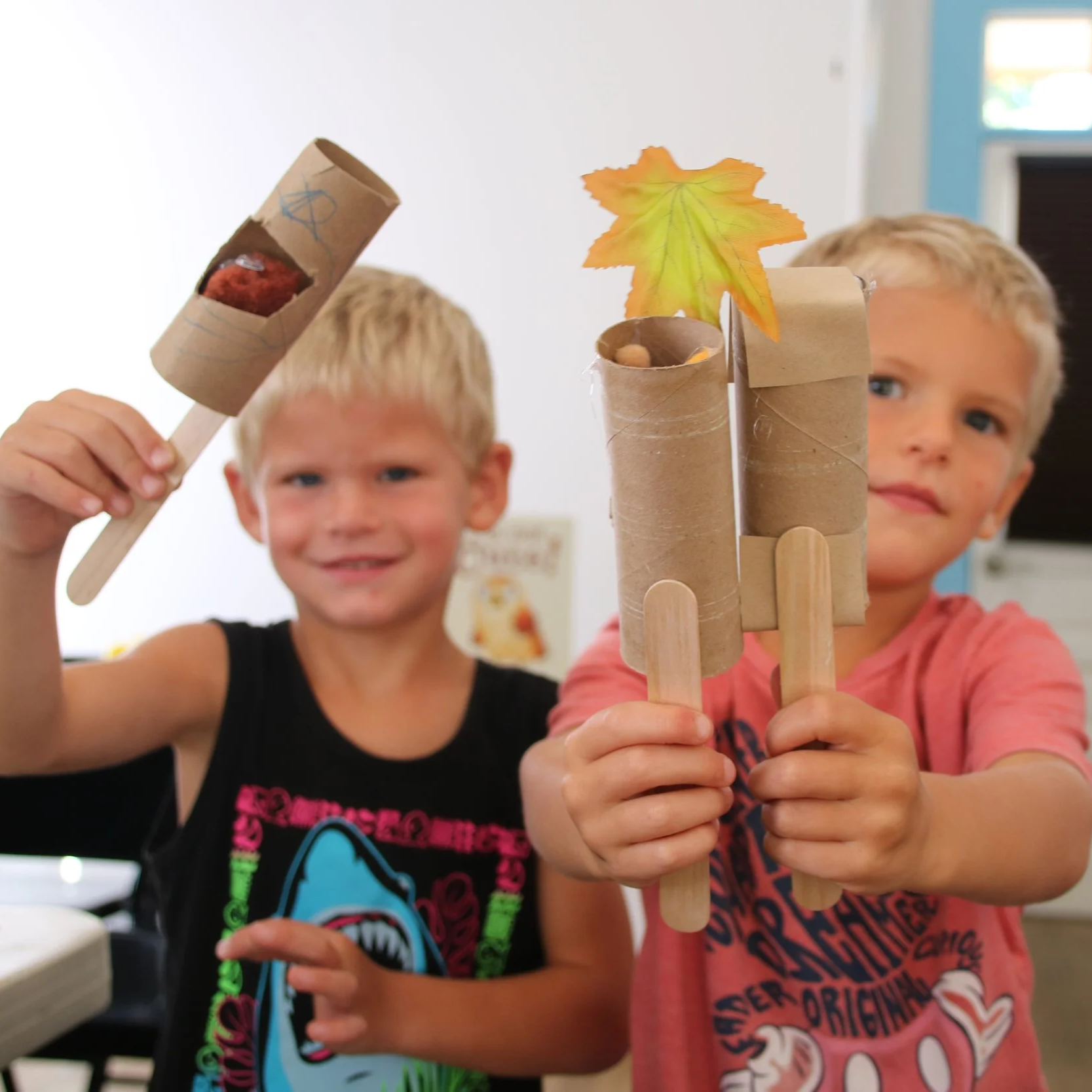Two young boys holding homemade marshmallow pops wrapped in paper tubes, decorated with colored leaves and sticks, in a classroom setting.