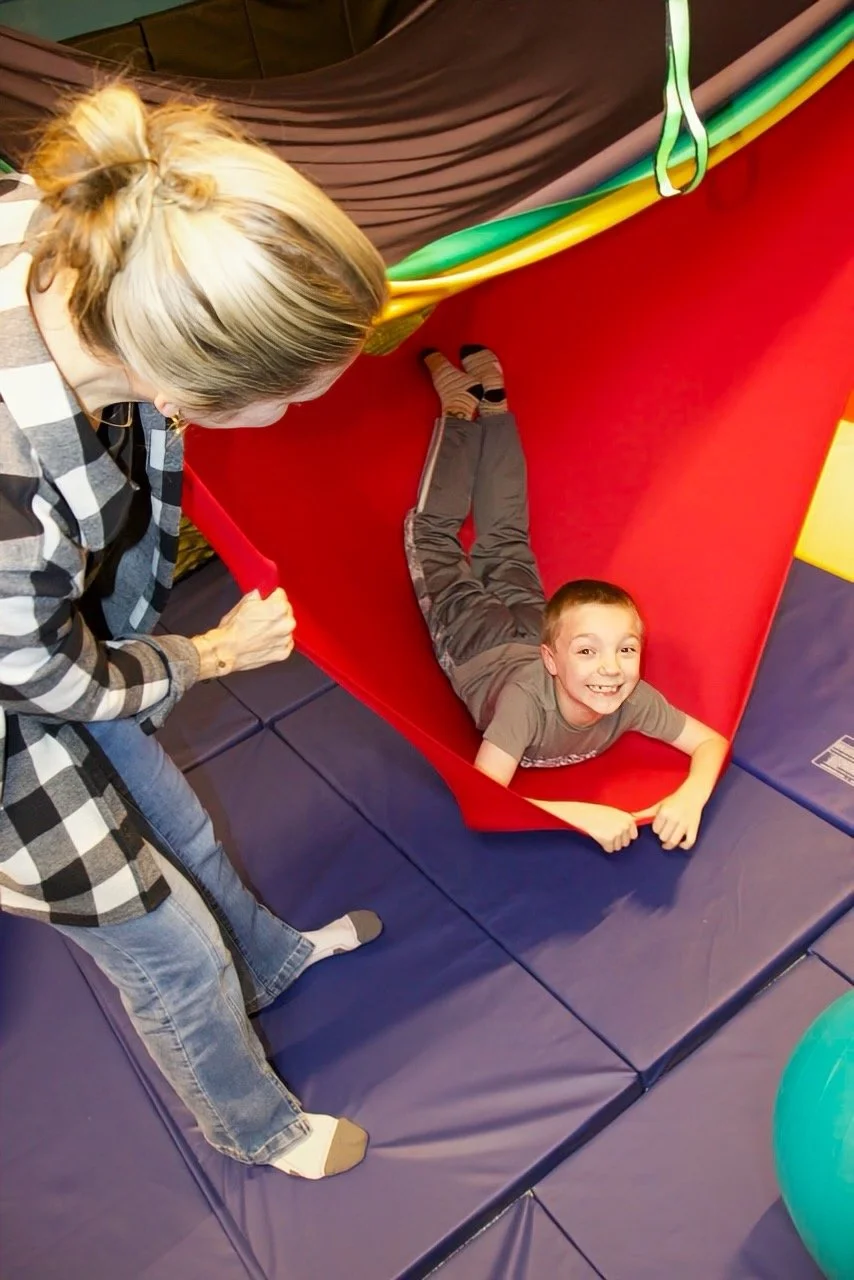 A young boy with a big smile lying at the bottom of a spandex swing with his occupational therapist standing near the edge.