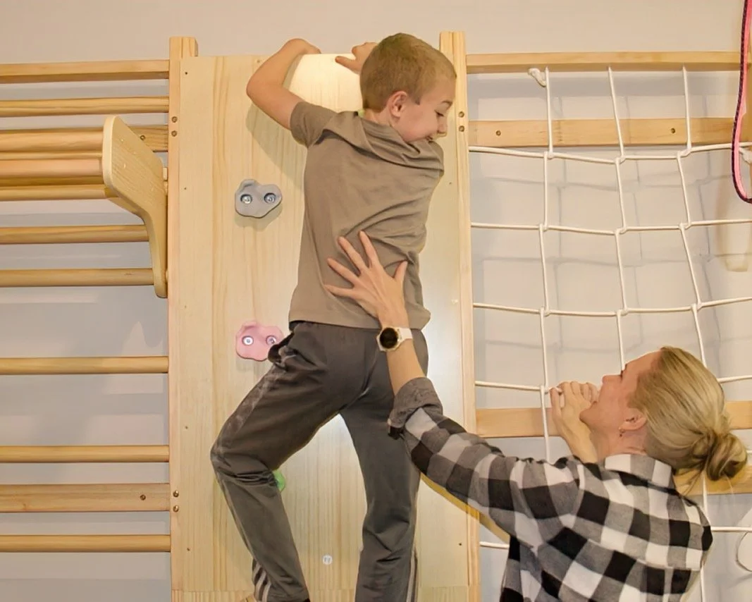An occupational therapist helping a young boy climb a wooden indoor climbing wall.