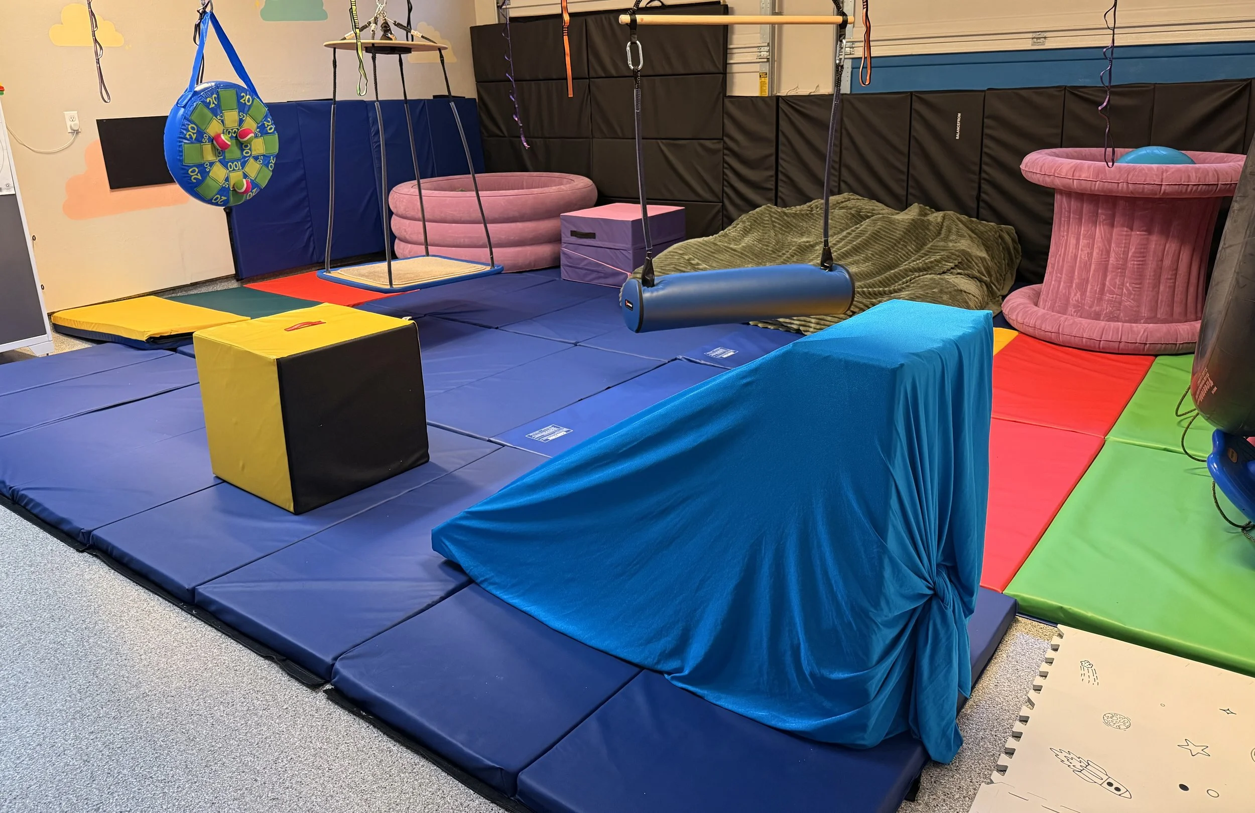 Indoor play area with padded flooring, hanging swings, foam climbing blocks, pink foam rings, and cushioned activity equipment.