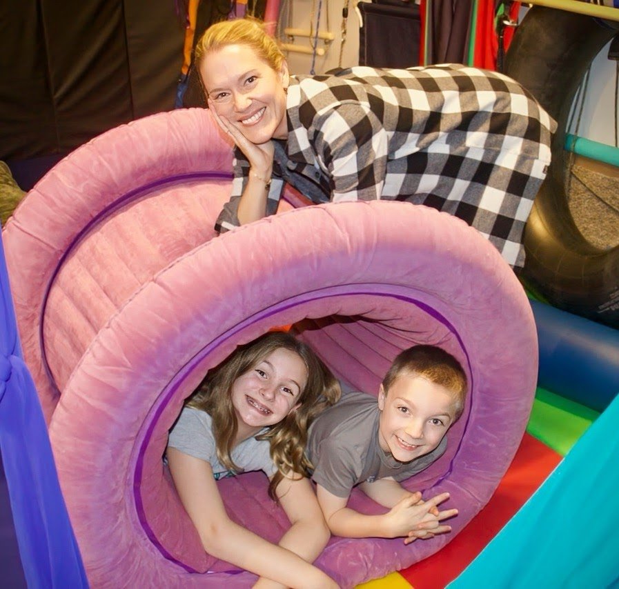 An occupational therapist and two children playing inside a large pink foam tube in a sensory integration clinic.