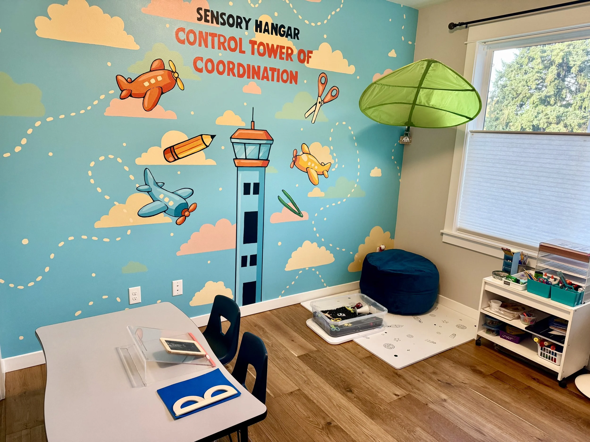 Children's playroom with a colorful wall mural featuring airplanes and clouds, labeled 'Sensory Hangar Control Tower of Coordination.' There is a table with chairs, a tablet, and a blue folder. The room has a window with blinds, a green hanging lamp, storage shelves with toys and art supplies, and a beanbag chair on the floor.