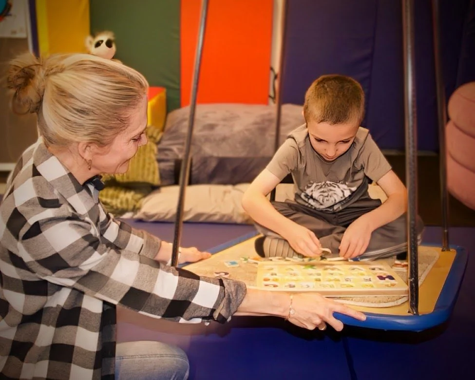 A woman with blonde hair tied back, wearing a black and white checkered shirt, is looking at a young boy sitting on a swing in a cozy room. The boy is focused on a board game or puzzle, engaged in playing. The room has colorful walls, a bed with pillows, and a stuffed panda toy in the background.