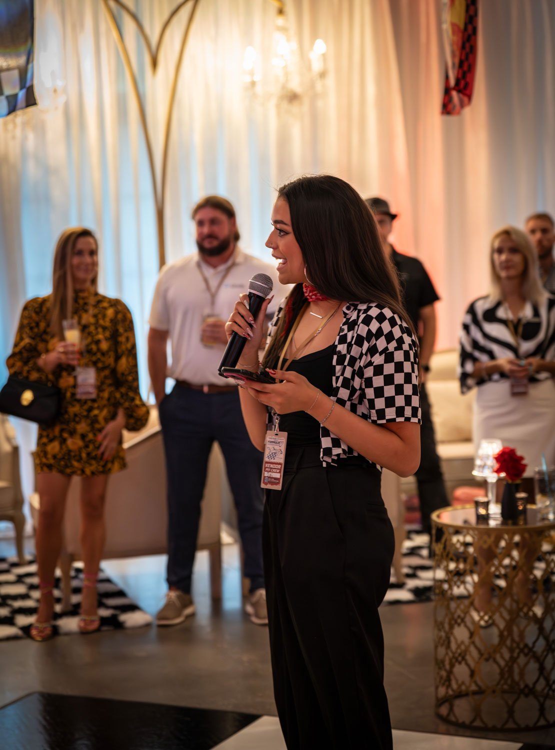 A woman with long dark hair, wearing a black outfit with a checkered short-sleeve jacket, speaking into a microphone at an indoor event with several people in the background.