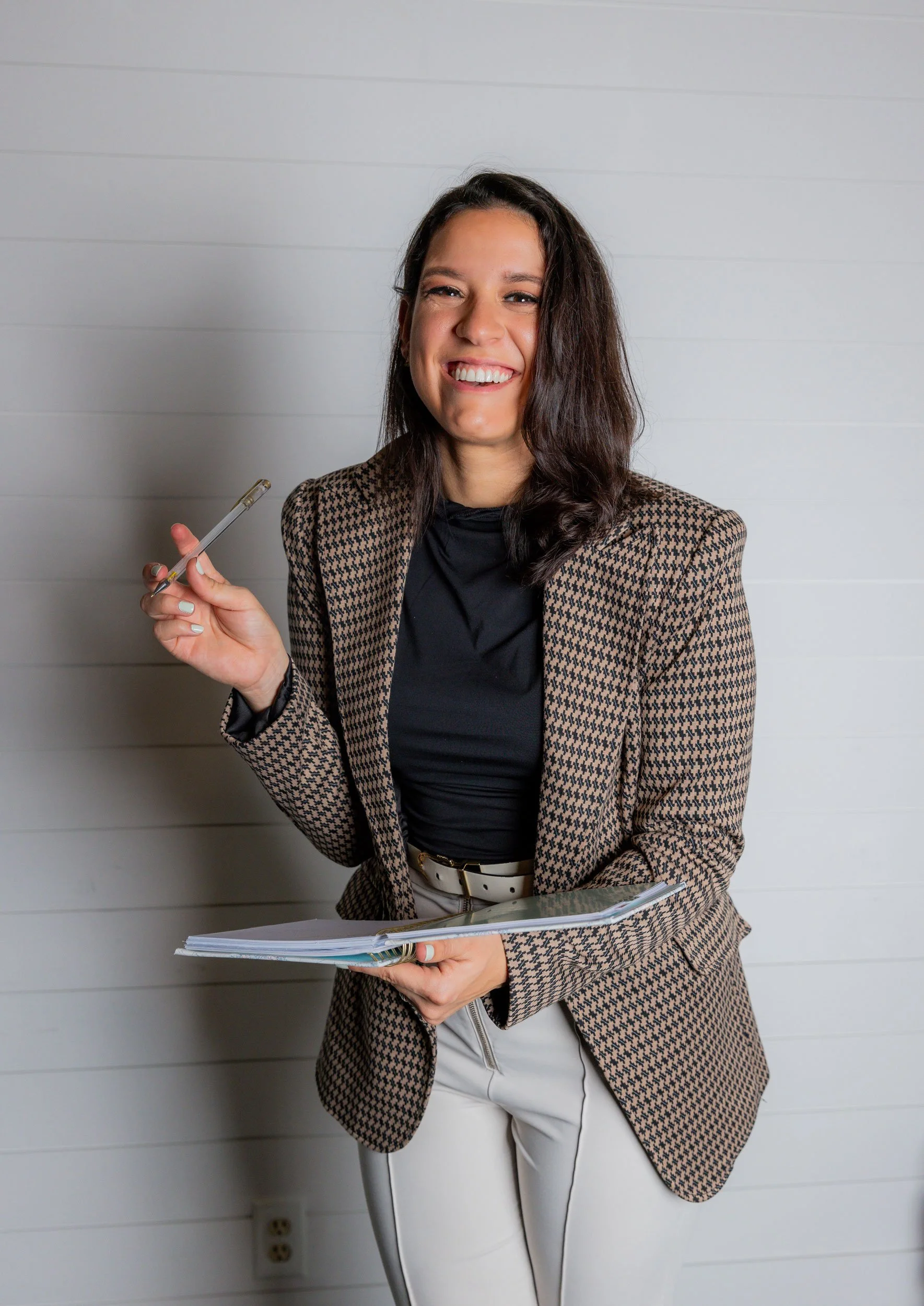 A woman smiling and holding a pen and a folder, wearing a patterned blazer and light-colored pants, standing against a white wall.