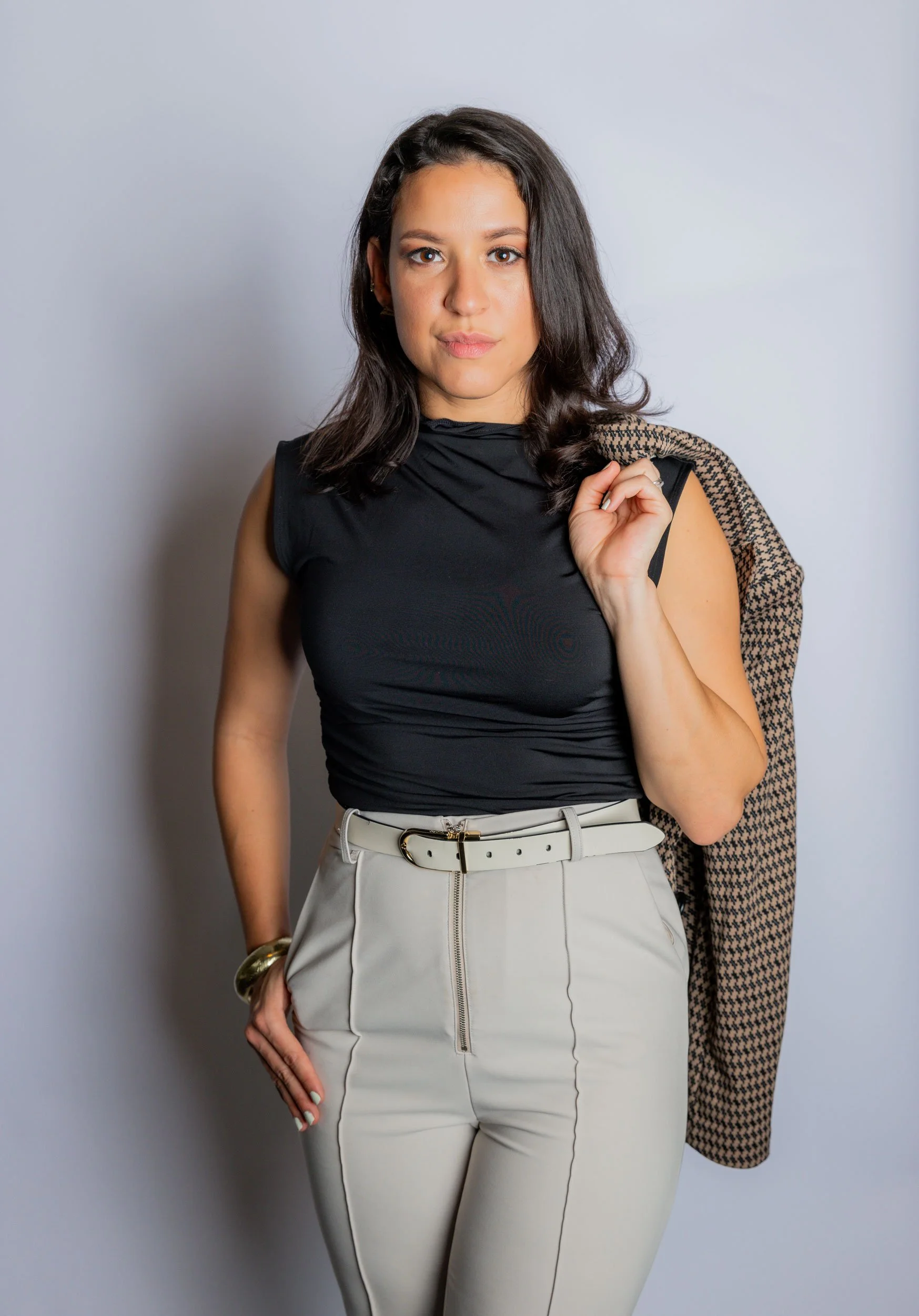 A woman with shoulder-length dark hair wearing a sleeveless black top, beige pants, and a houndstooth blazer draped over her shoulder holding a bag, standing against a light-colored background.